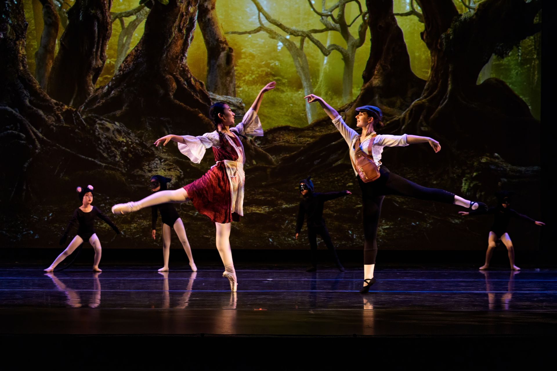 Ballet dancers in red and white costumes perform onstage beneath a forest backdrop.