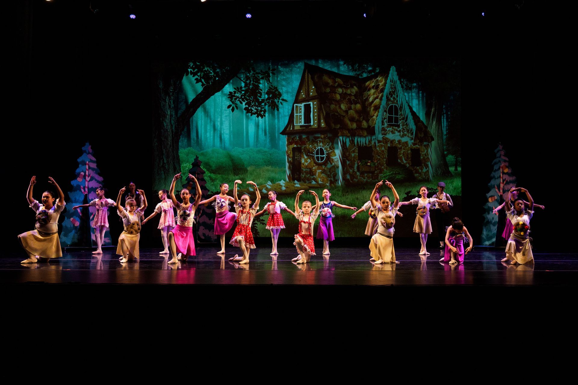 Children in colorful costumes performing on a lit stage with a painted haunted house backdrop
