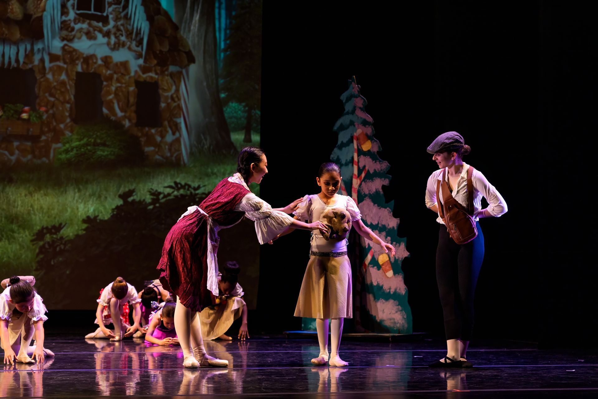 Three dancers onstage in period costumes, with two seated figures and a painted forest backdrop.