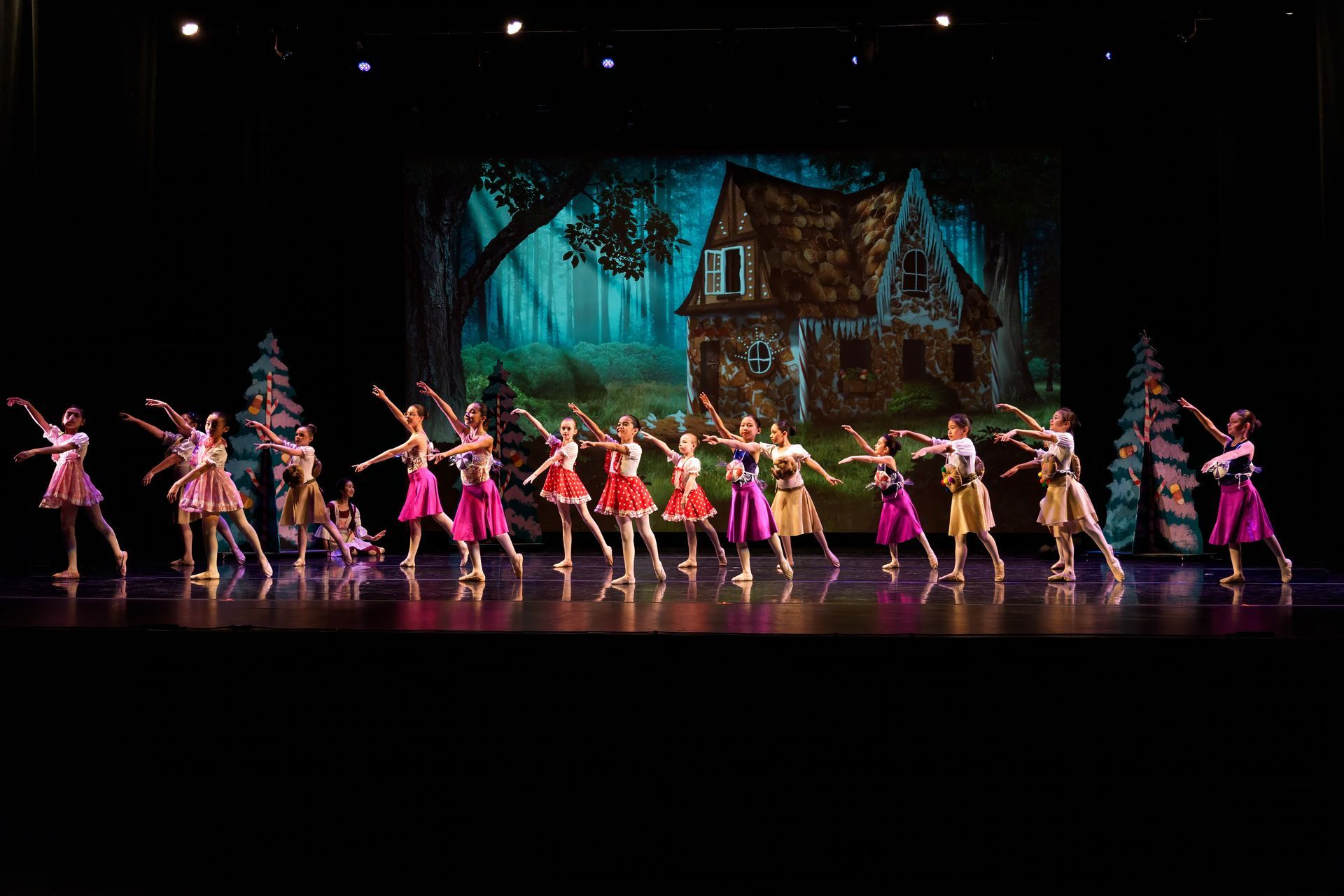 Ballet troupe in pink costumes onstage before a painted house backdrop, arms raised in a synchronized pose