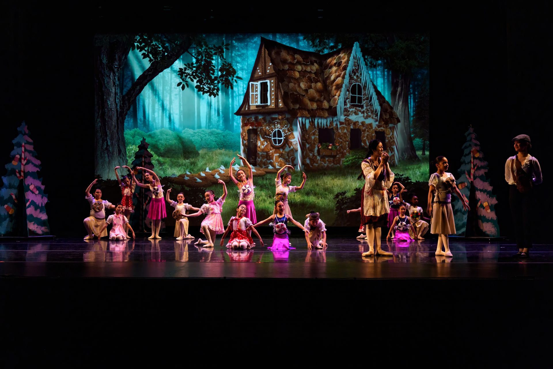A stage performance with dancers in colorful costumes in front of a glowing cottage and forest backdrop