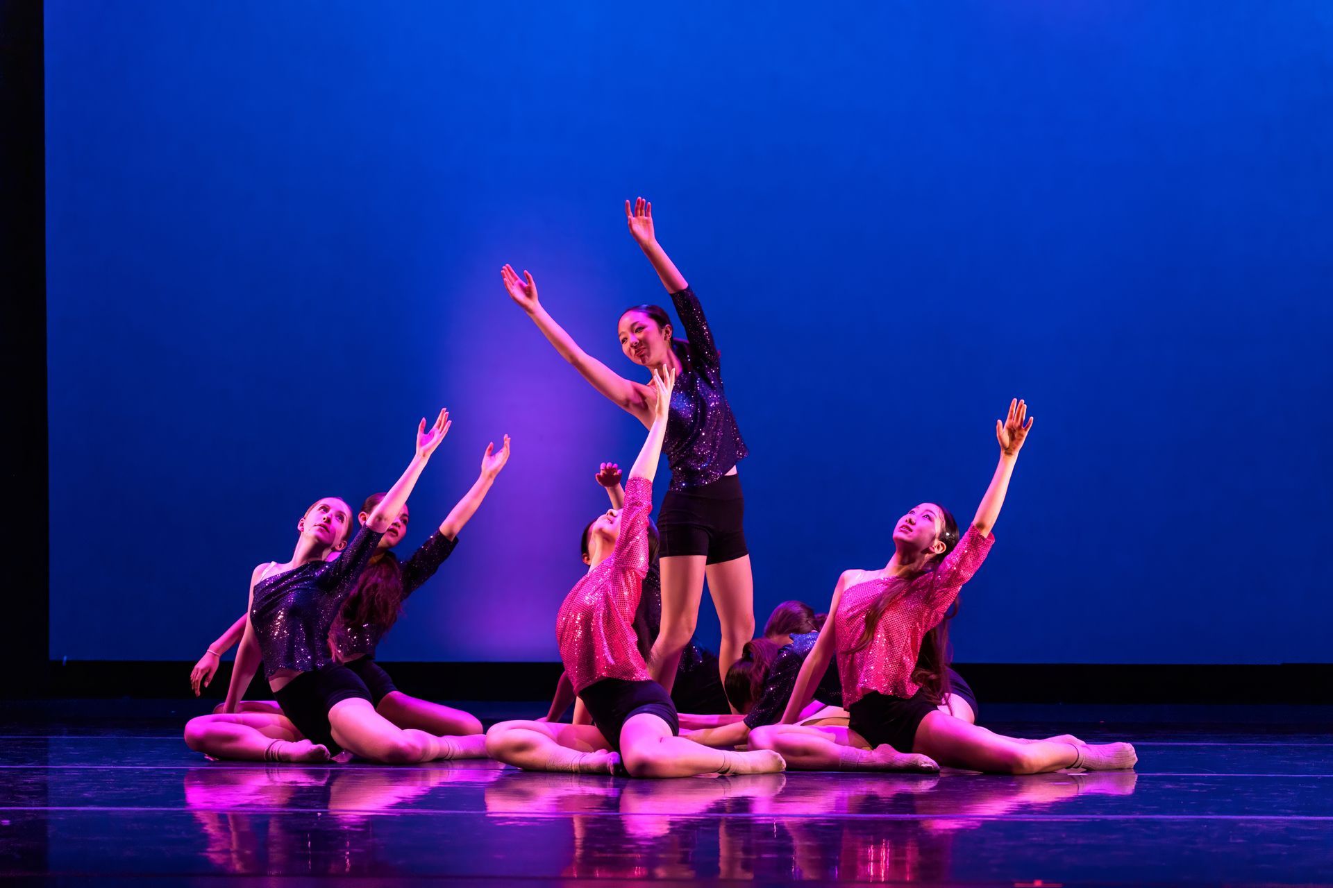 Three dancers in pink and black pose on a blue-lit stage, arms raised in a dramatic formation