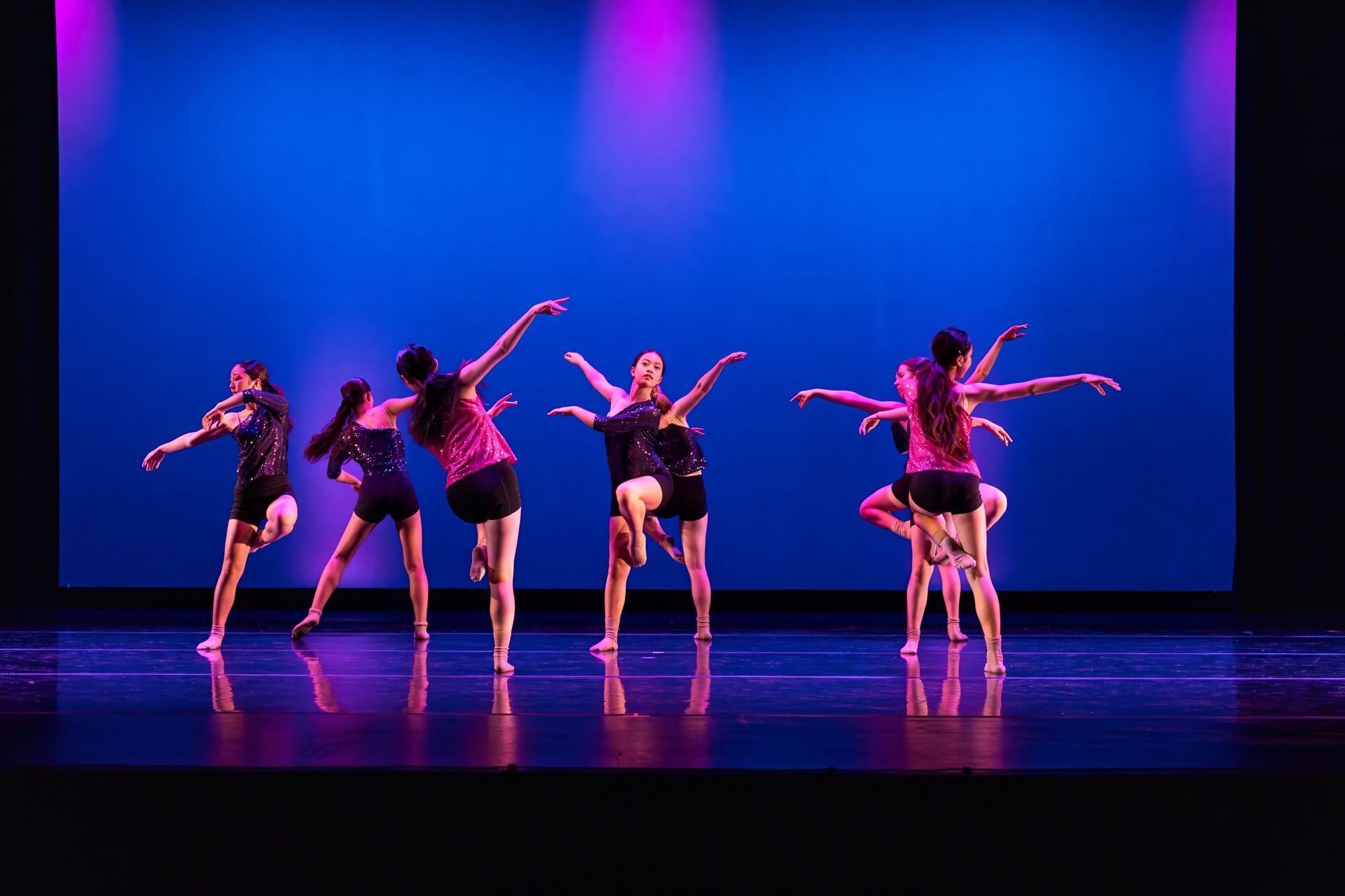 Six dancers perform on a blue-lit stage with pink lighting and reflected floor.