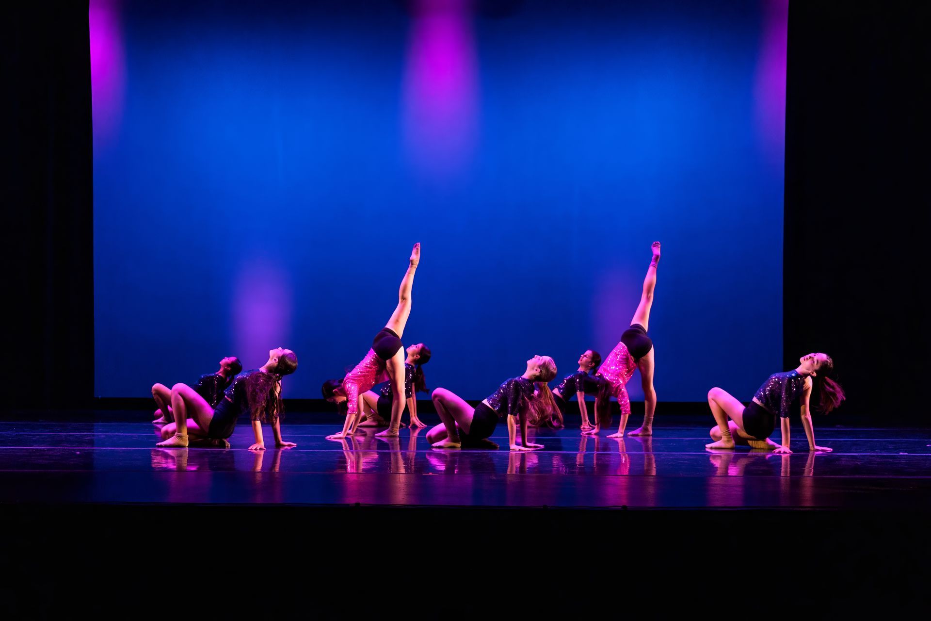 Dance troupe performing on a blue-purple lit stage, with dancers posed low and one leg raised vertically.