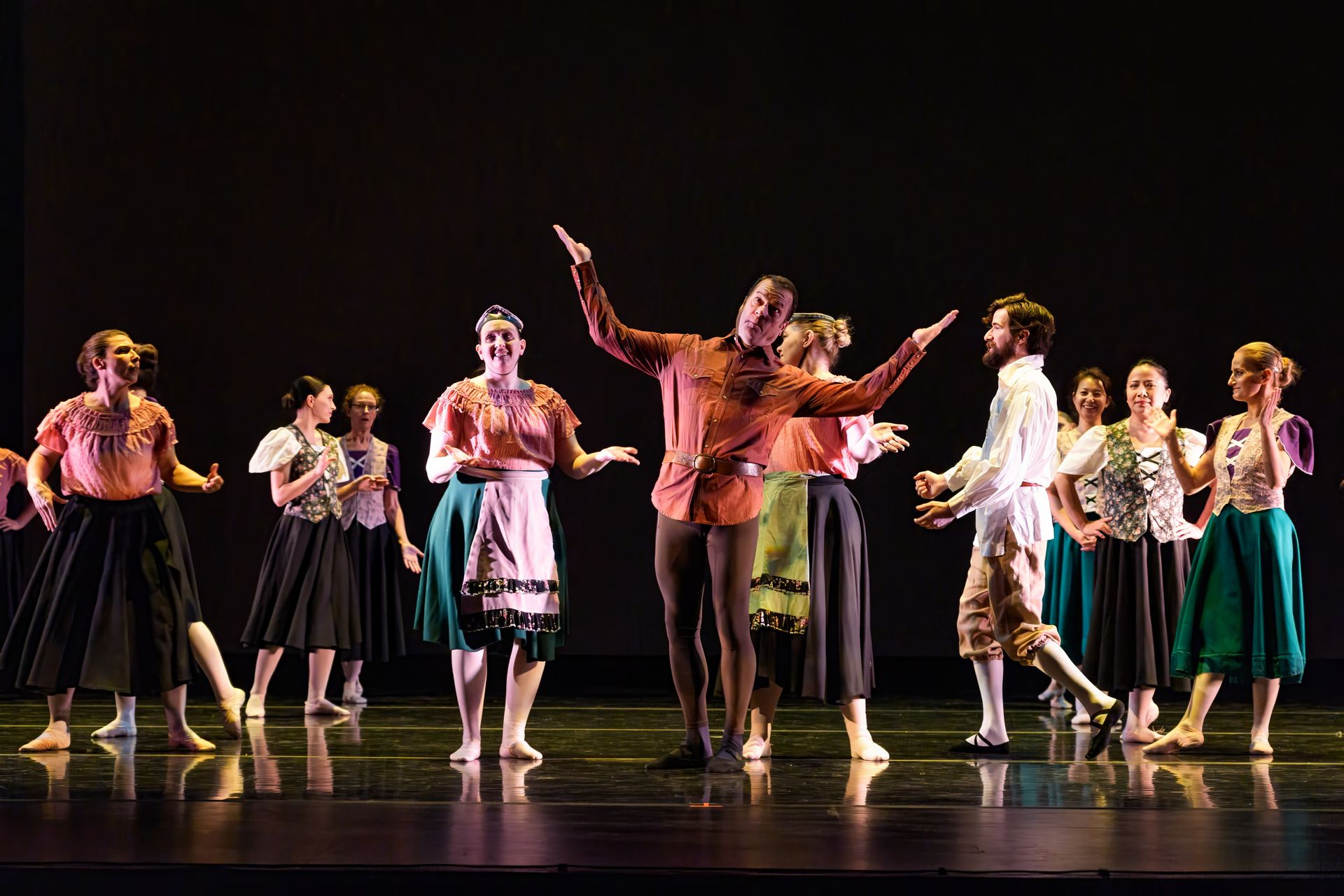 Dancers on a dark stage, one in a red shirt center with arms raised, surrounded by a semicircle of performers