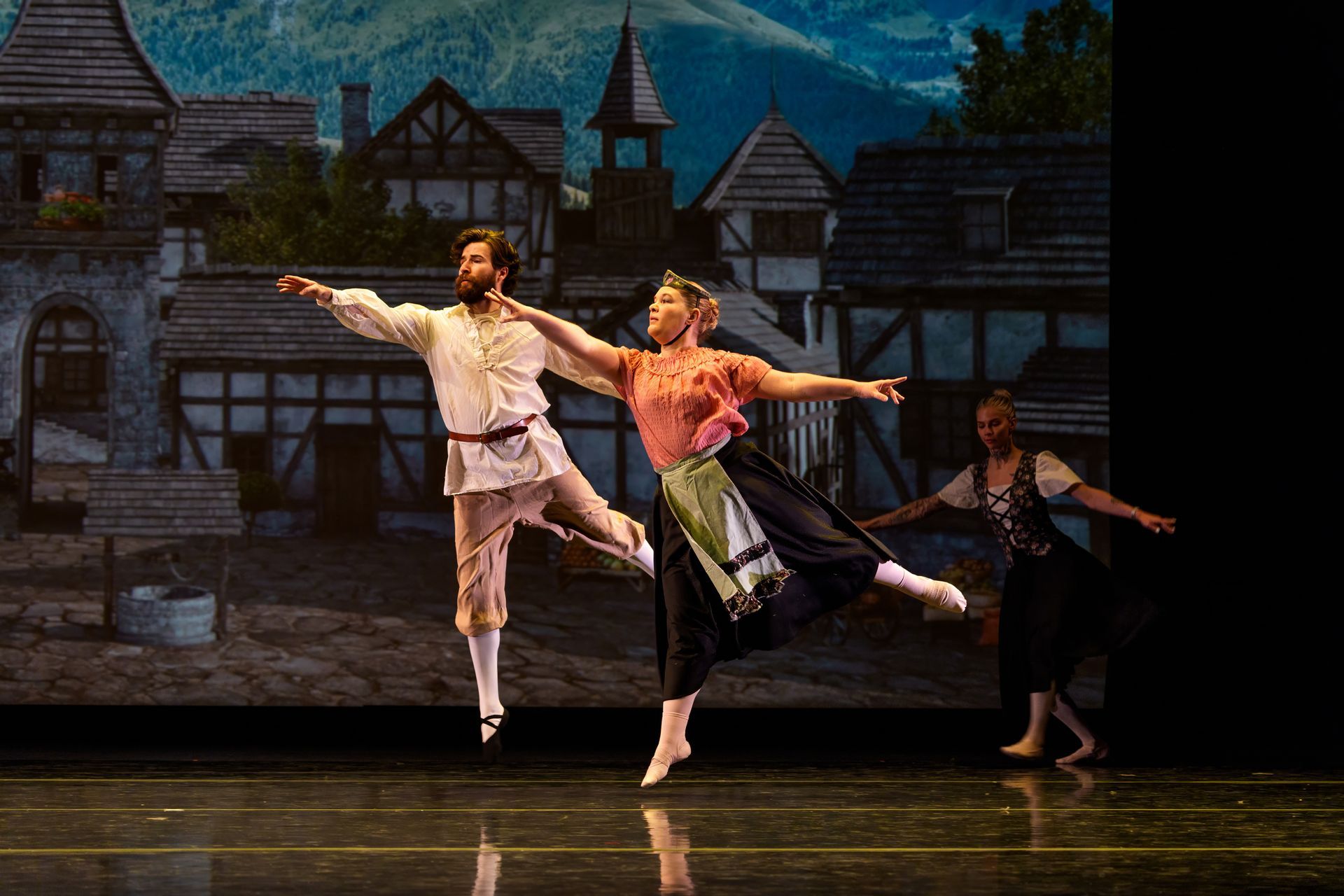 Three dancers pose mid-leap on a stage with a village backdrop under warm lights