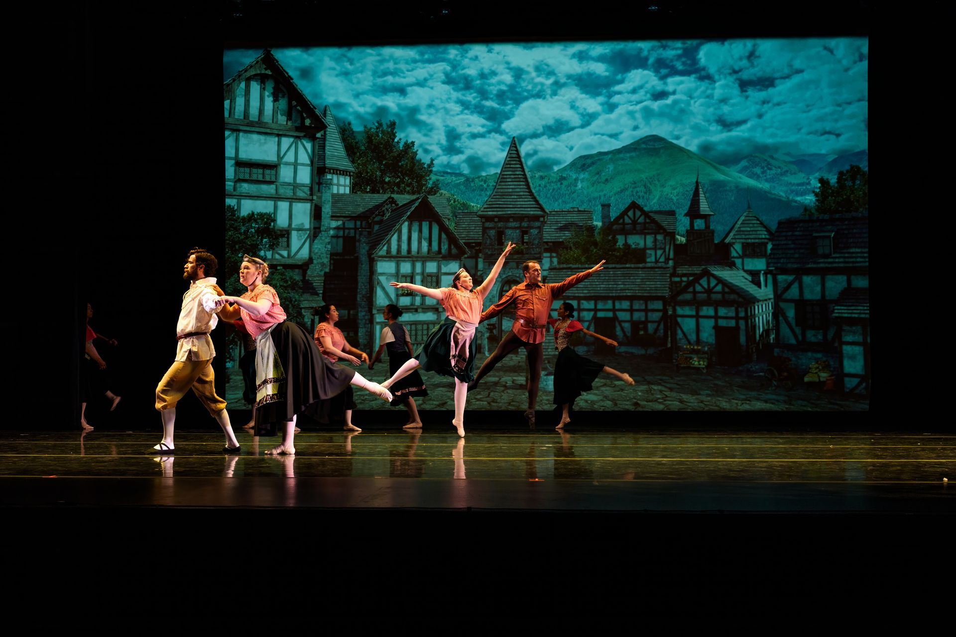 Dancers perform on a stage before a painted village backdrop with teal lighting.