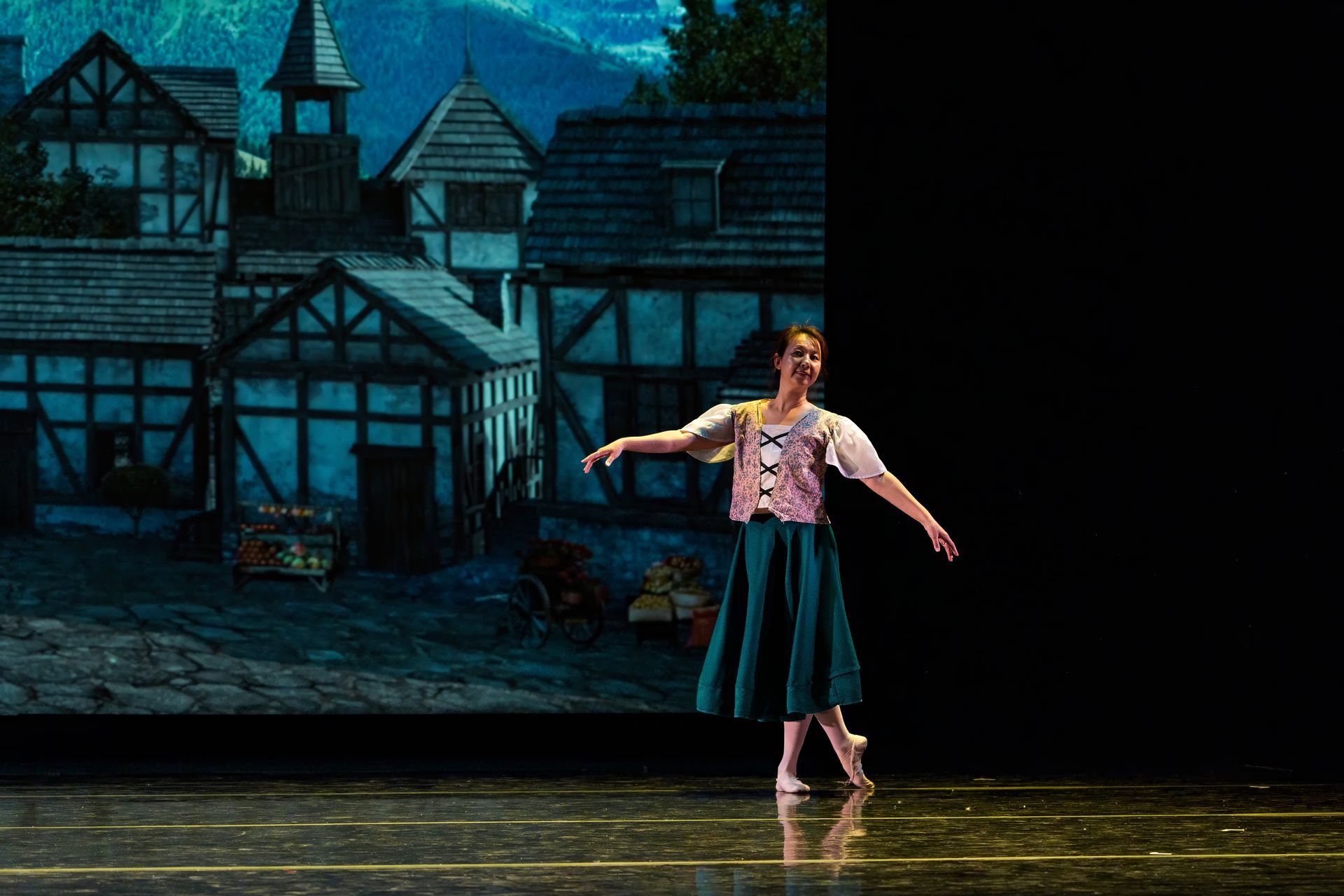 Ballet dancer in a pink costume stands onstage before a painted village backdrop.