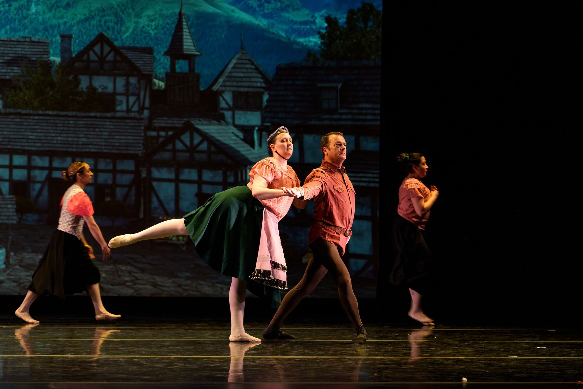 Ballet dancers performing onstage in front of a painted village backdrop, with one dancer held horizontally.