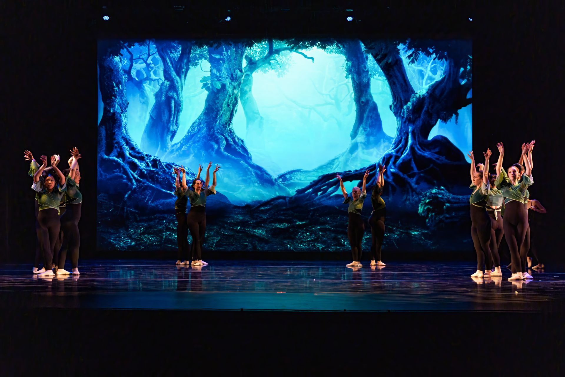 Four dancers on a stage in front of a glowing blue forest backdrop with raised arms