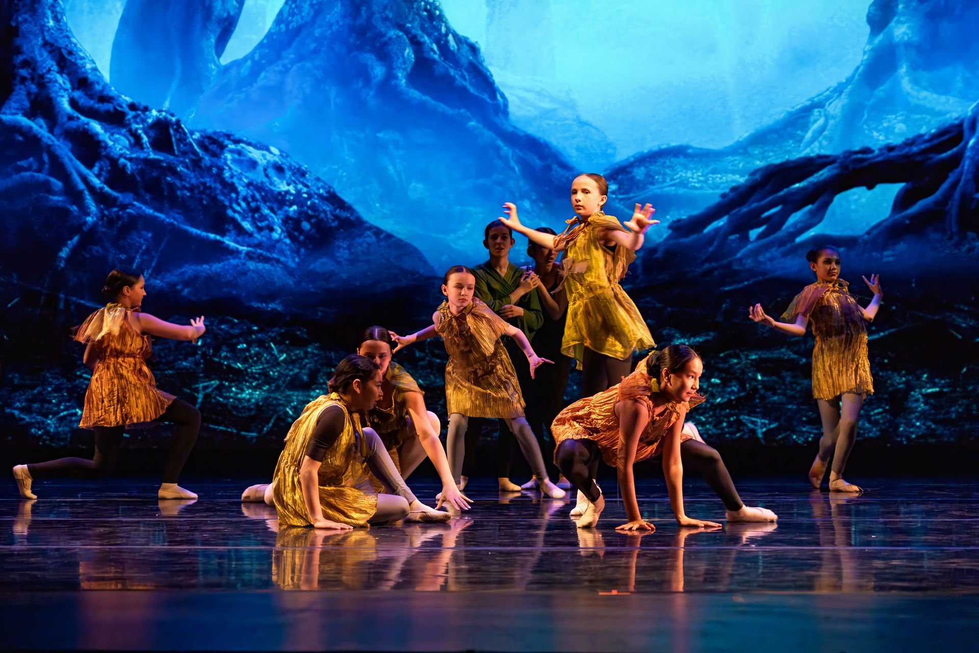 Dancers in yellow costumes perform on a blue-lit stage with a dramatic mountain backdrop.