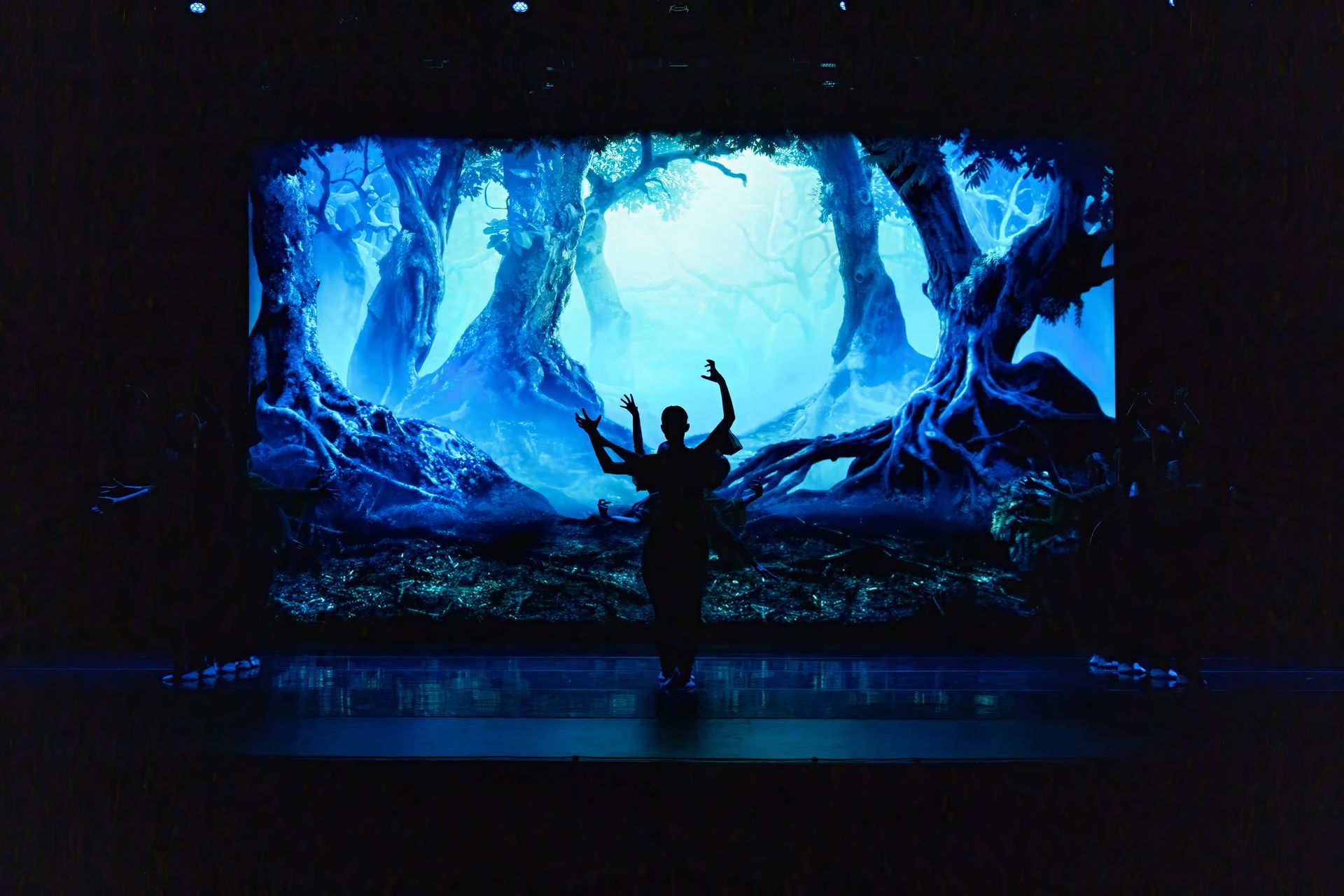 Silhouetted person with raised arms in a glowing blue forest scene on a large screen