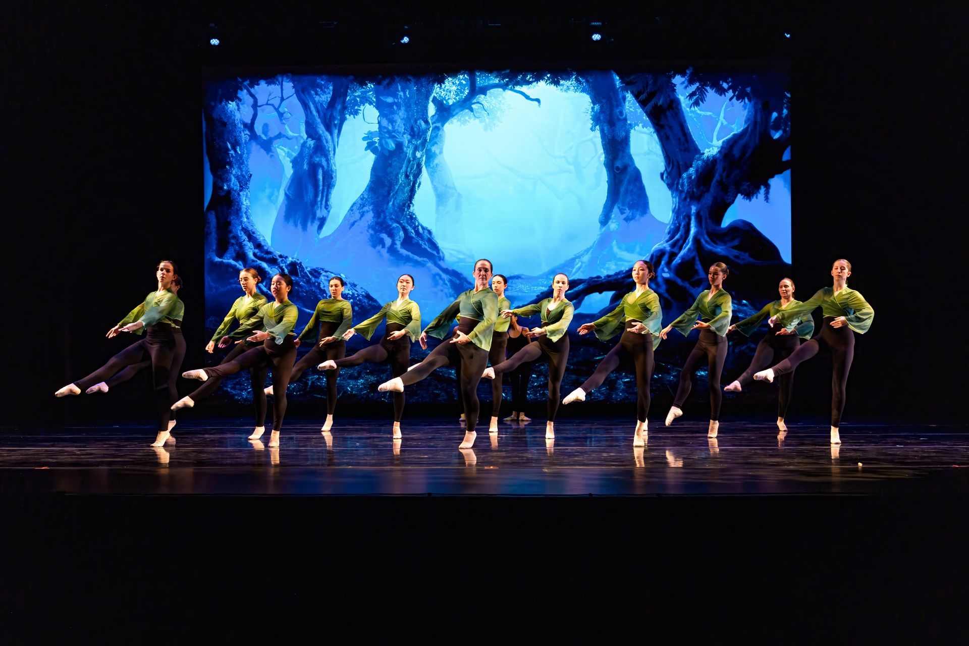 Dancers in green costumes perform a synchronized routine on a stage with a blue forest backdrop.