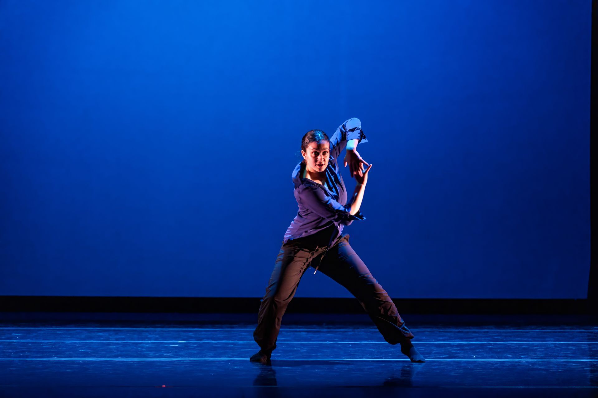 Dancer in dark clothes performing on a blue-lit stage against a plain blue backdrop
