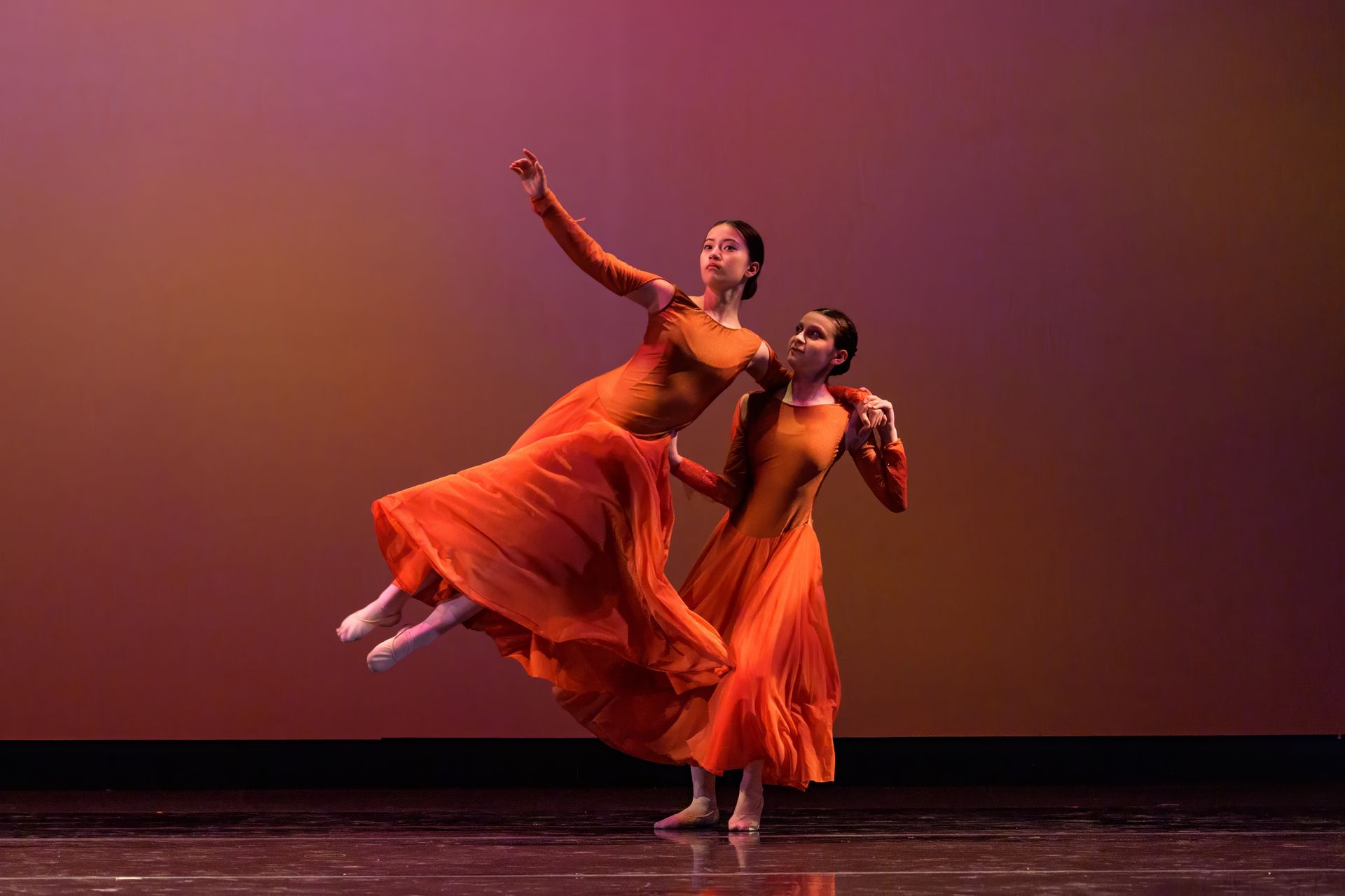 Two dancers in flowing orange dresses perform a ballet pose on a stage with a purple backdrop