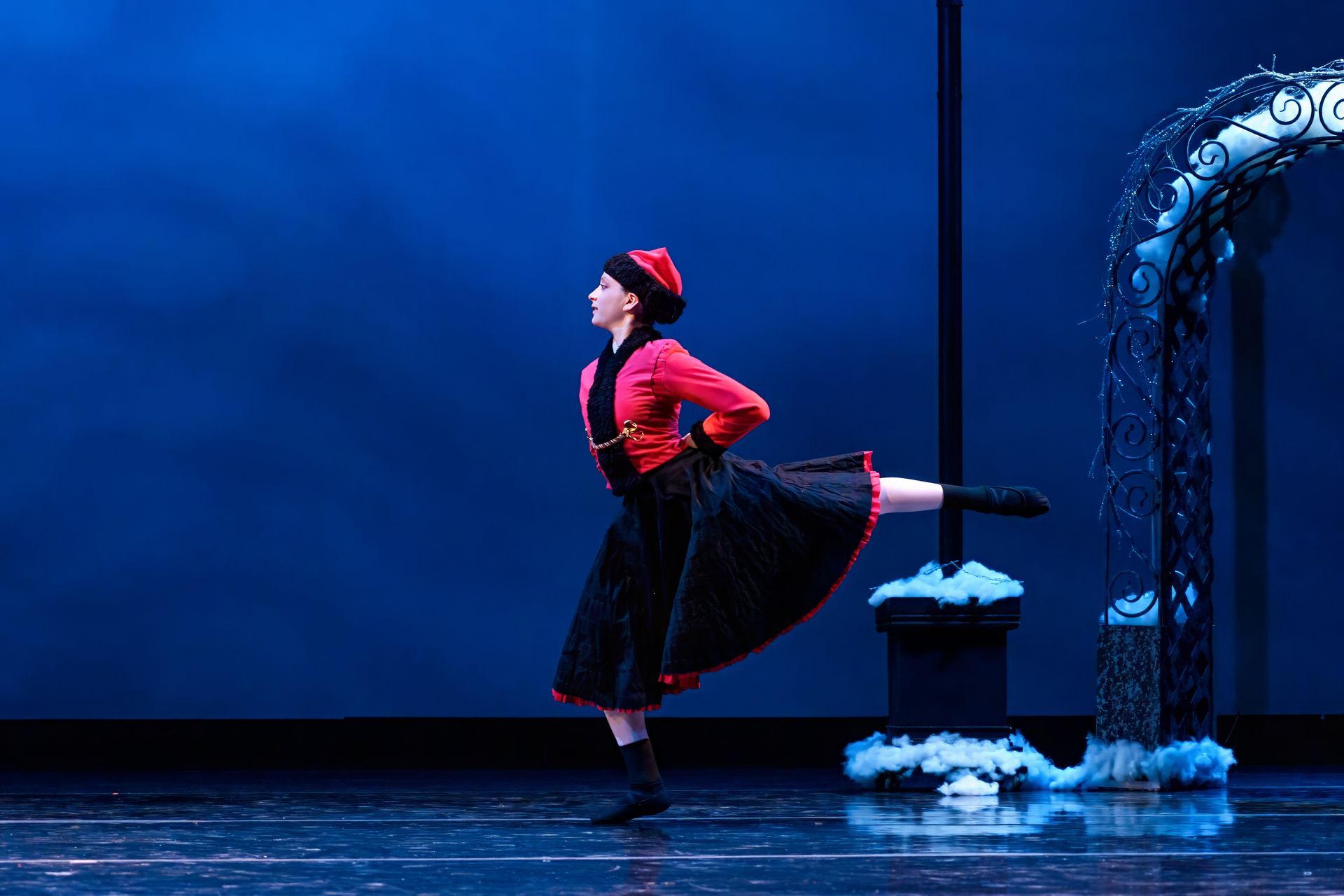 A dancer in red and black stands on one leg beside a blue-lit stage arch with snow-like props.