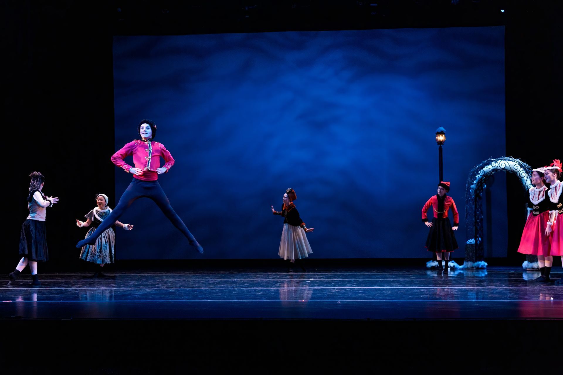 Performers on a blue-lit stage mid-dance, wearing colorful costumes with one dancer leaping high.