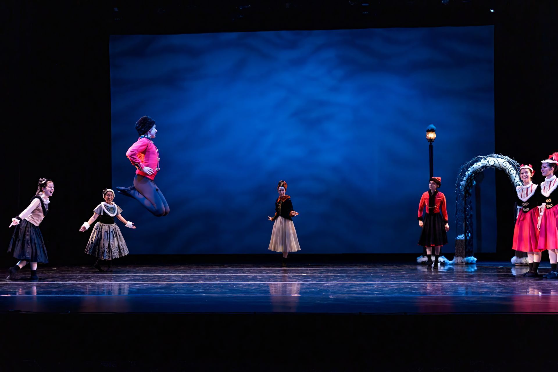 Dancers in colorful costumes performing on a dimly lit stage with a blue backdrop