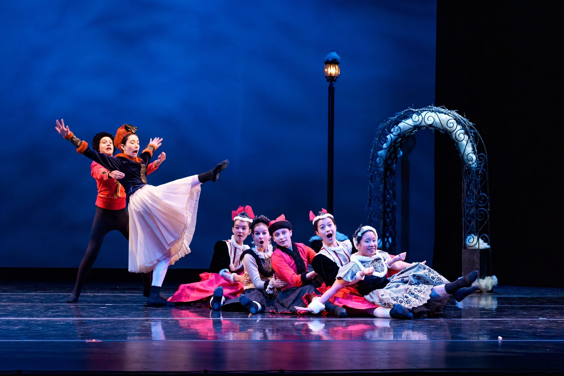 Ballet dancers in red and white perform onstage with a blue backdrop and glowing arch.
