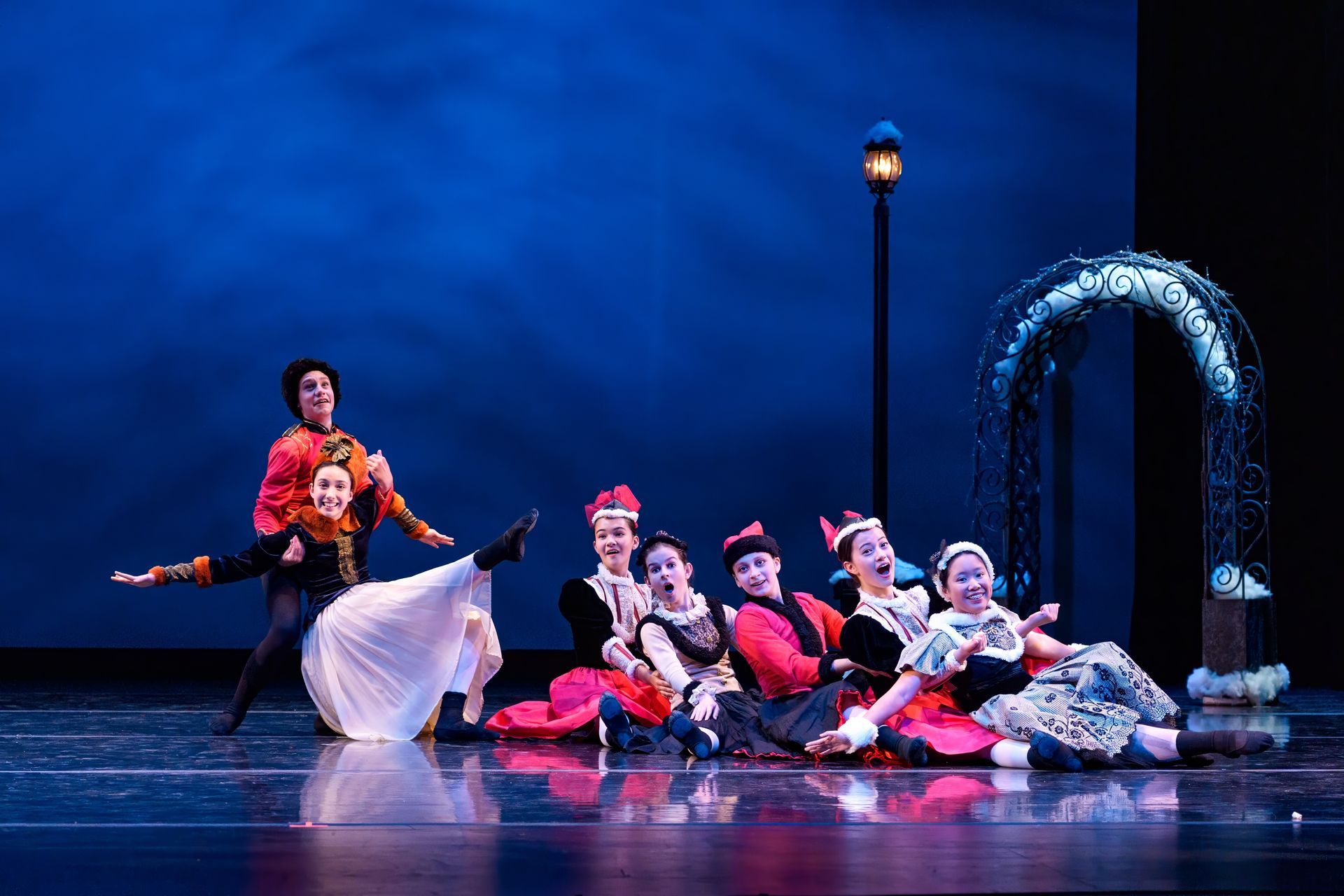 Ballet dancers performing on a blue-lit stage with a lamppost and arched prop, one dancer in white at left.