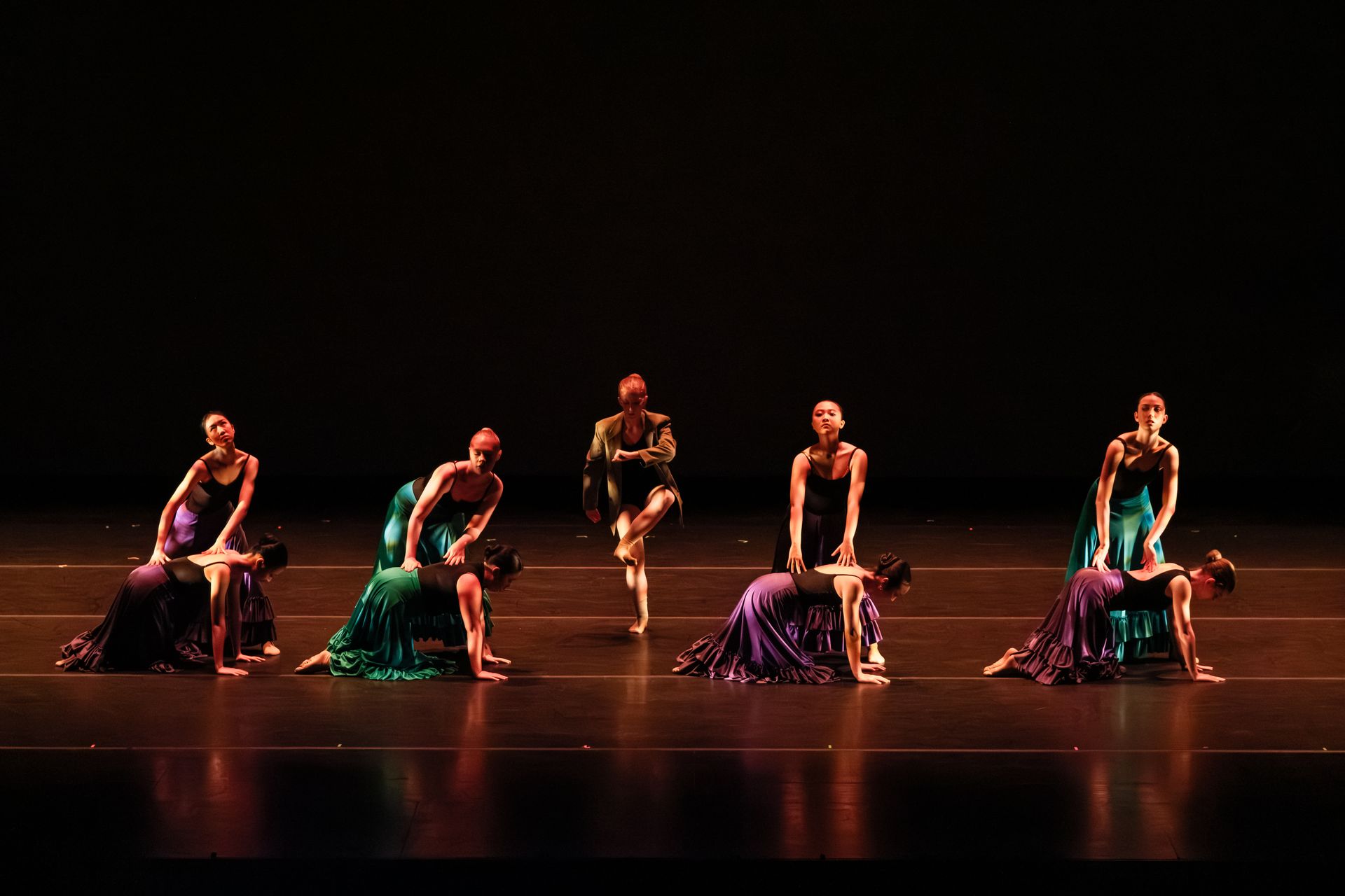 Dancers in green and purple costumes pose on a dark stage under warm spotlights.