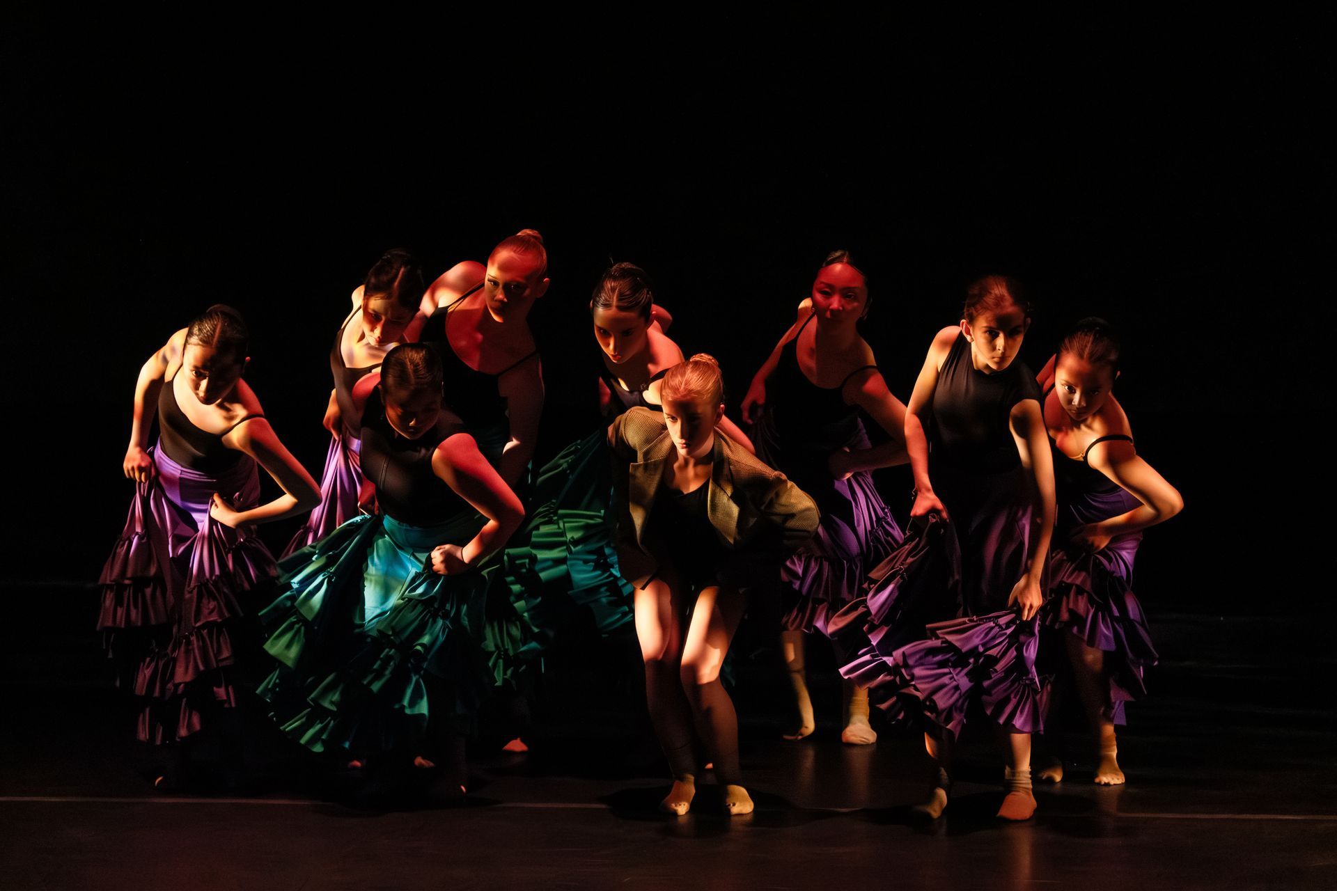 Group of dancers performing on a dark stage in colorful costumes, mid-routine under warm spotlight