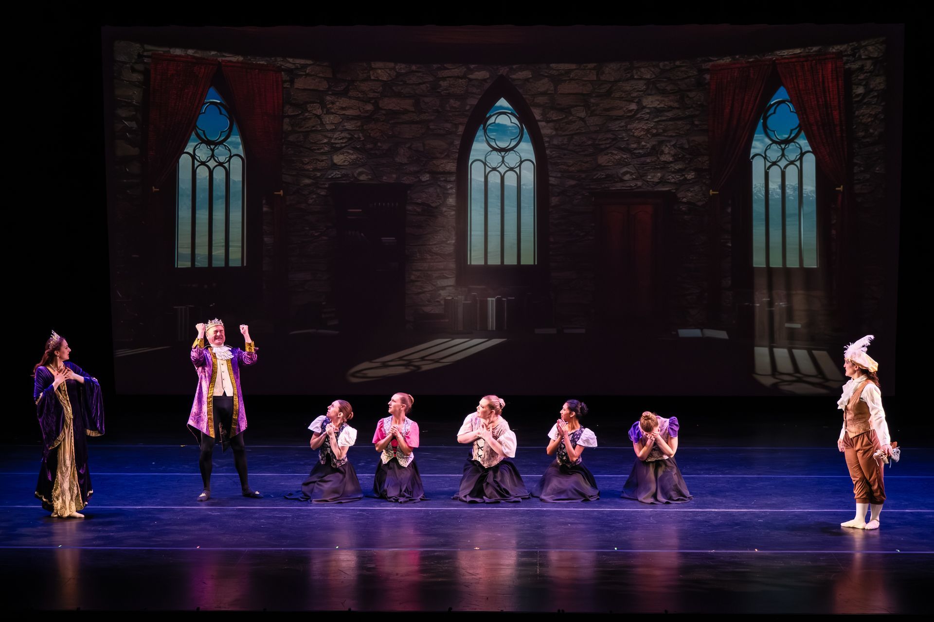 Dancers performing on a stage with a stone-wall backdrop and three tall stained-glass windows