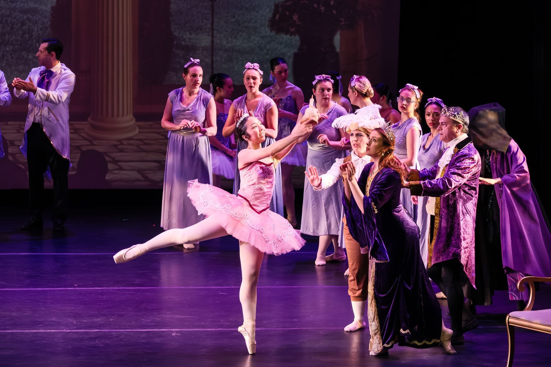 Ballet dancer in pink tutu poses en pointe onstage beside a line of costumed performers watching