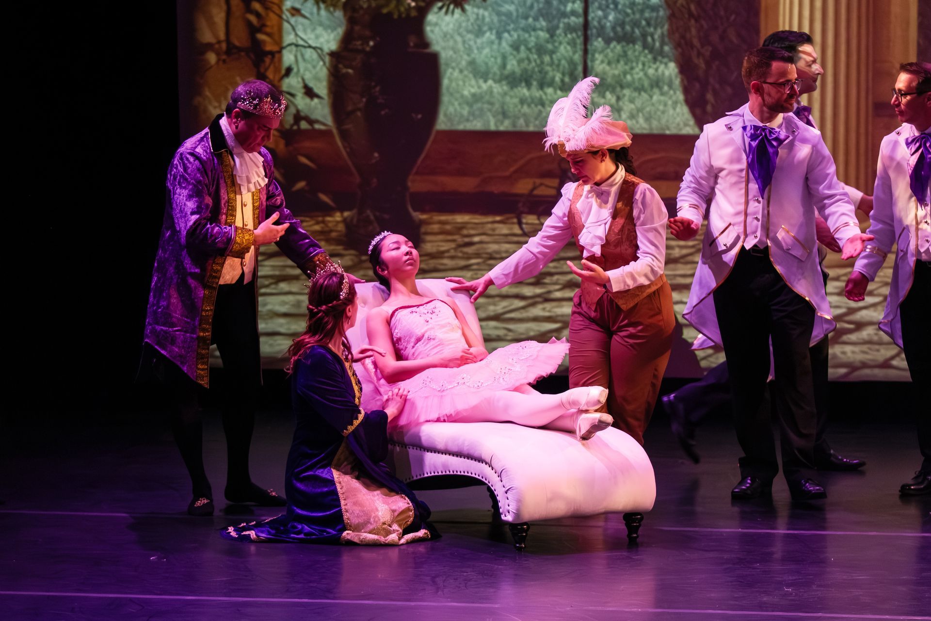 Stage performance with a person reclining on a white chaise while four costumed actors stand around them under purple lighting
