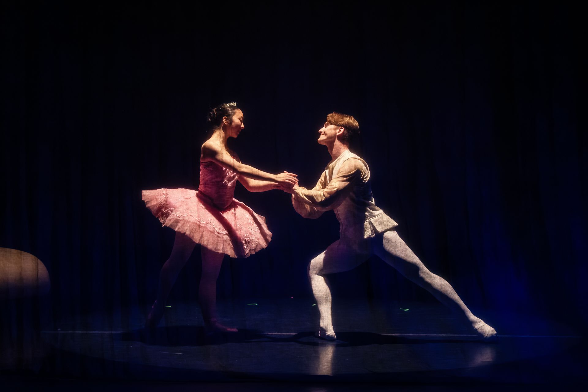 Two ballet dancers in pink and white costumes hold hands in a dramatic stage pose under spotlight.