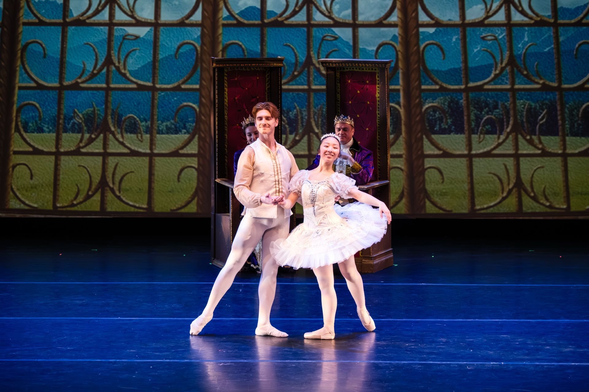 Two ballet dancers in white costumes pose on a blue stage before ornate green windows.