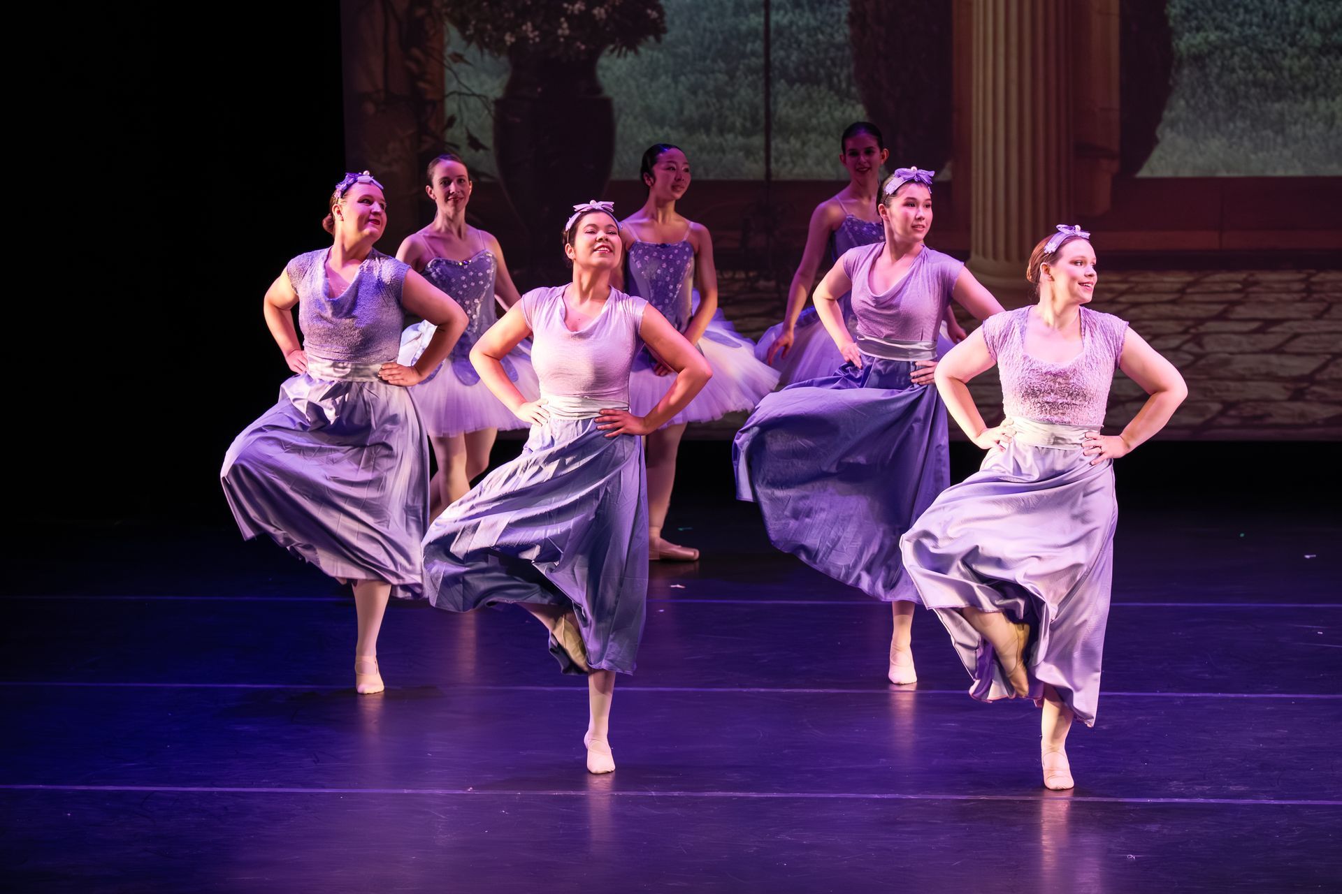 Dance troupe in lavender costumes performing synchronized kicks on a stage with purple lighting