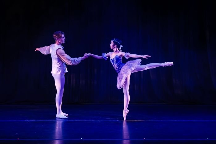 Two ballet dancers in white costumes performing an arabesque on a dark blue stage under purple lighting