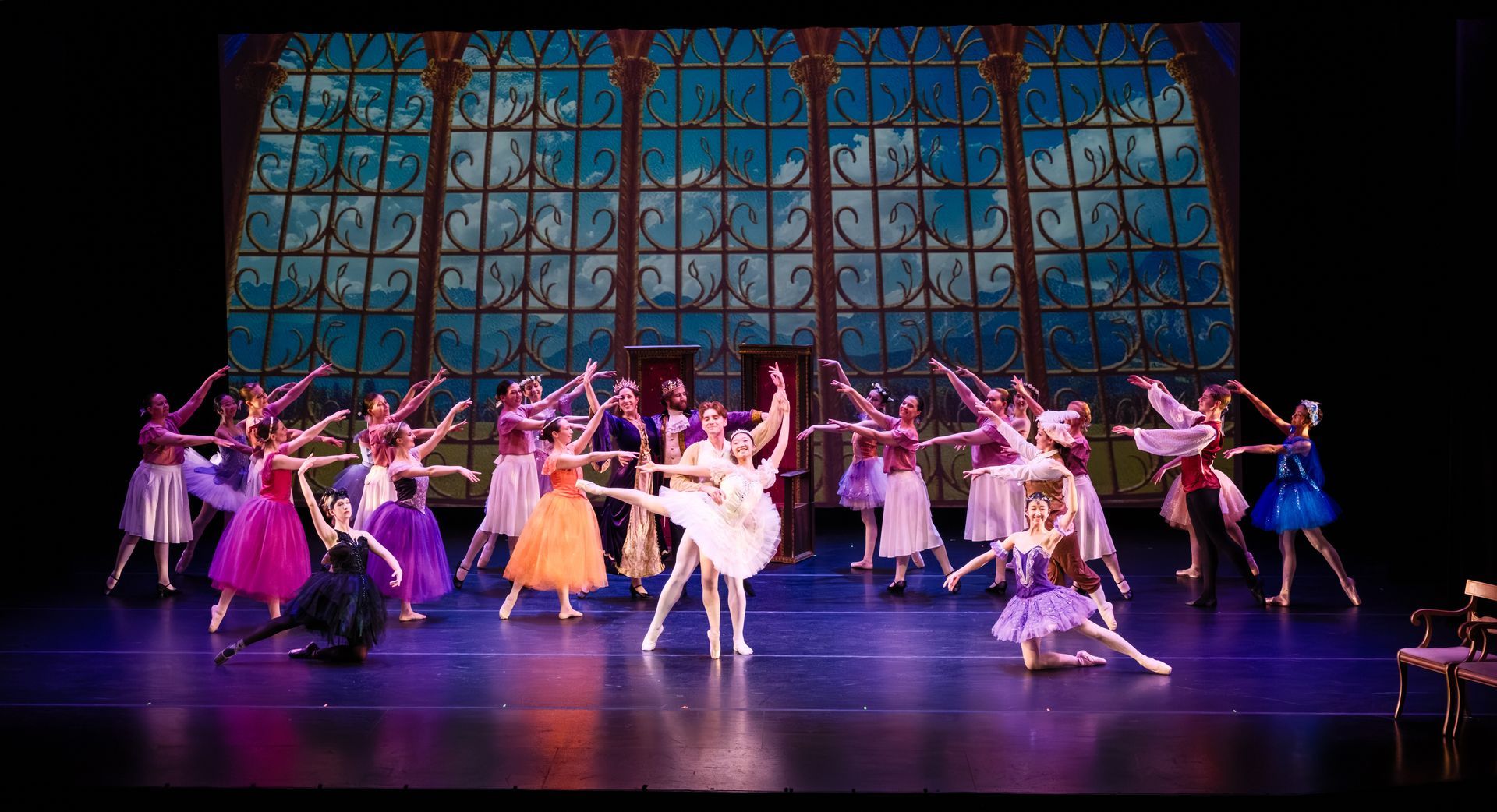 Ballet troupe performing onstage in colorful costumes before a stained-glass backdrop