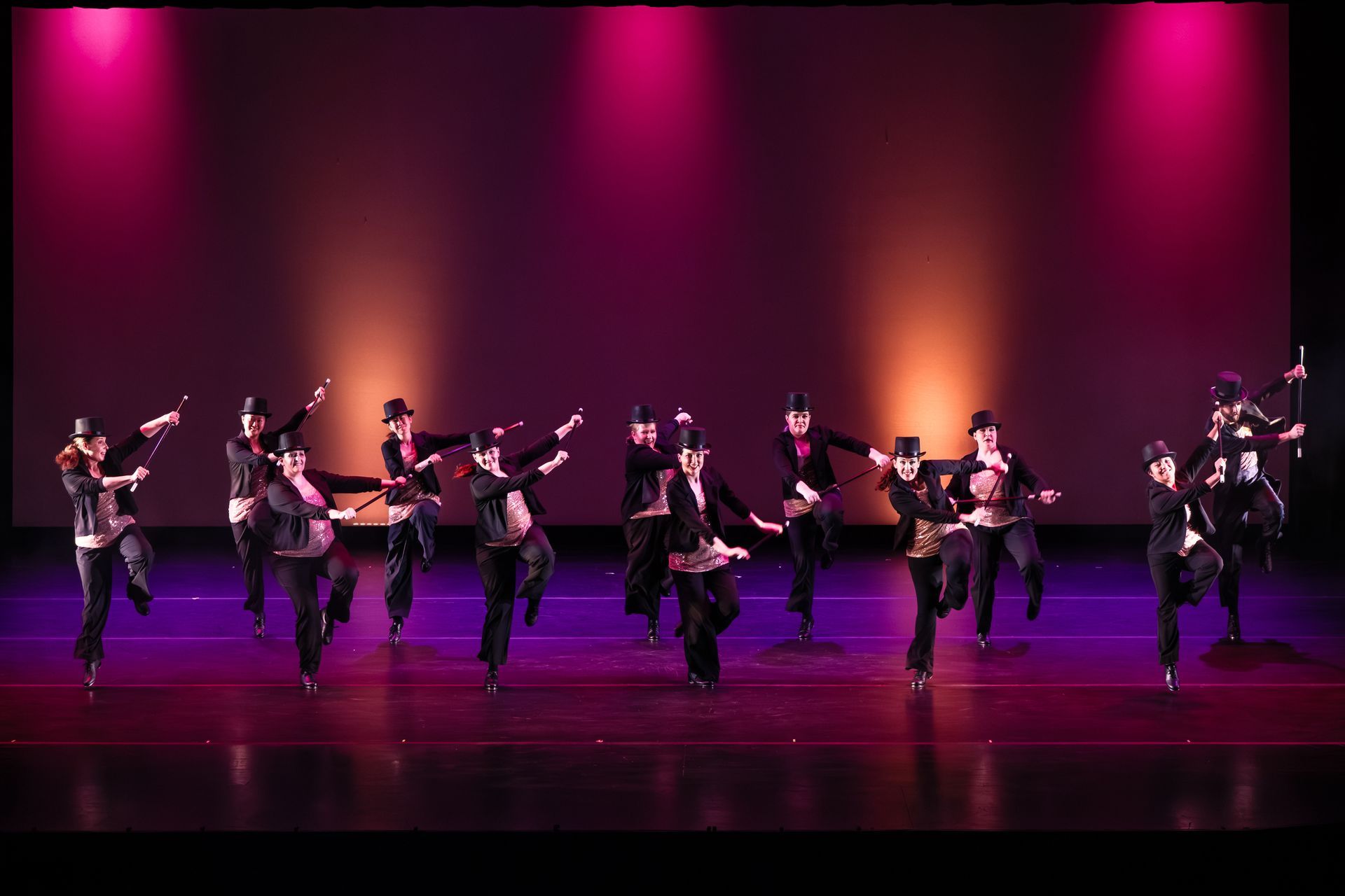 Line of dancers in black costumes performing on a stage lit with pink and purple lights
