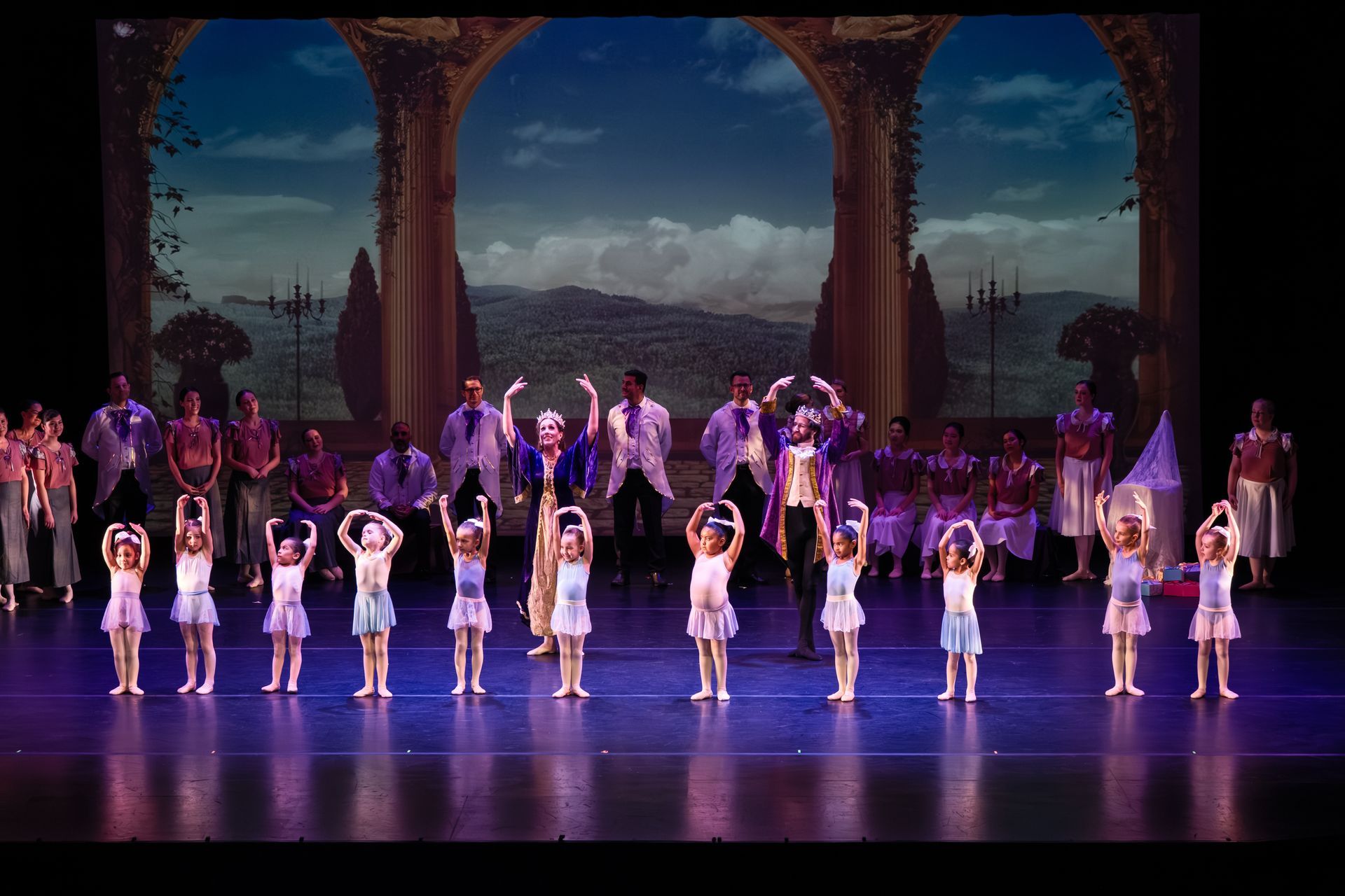 Ballet ensemble on a theater stage with pink and purple costumes under ornate arches.