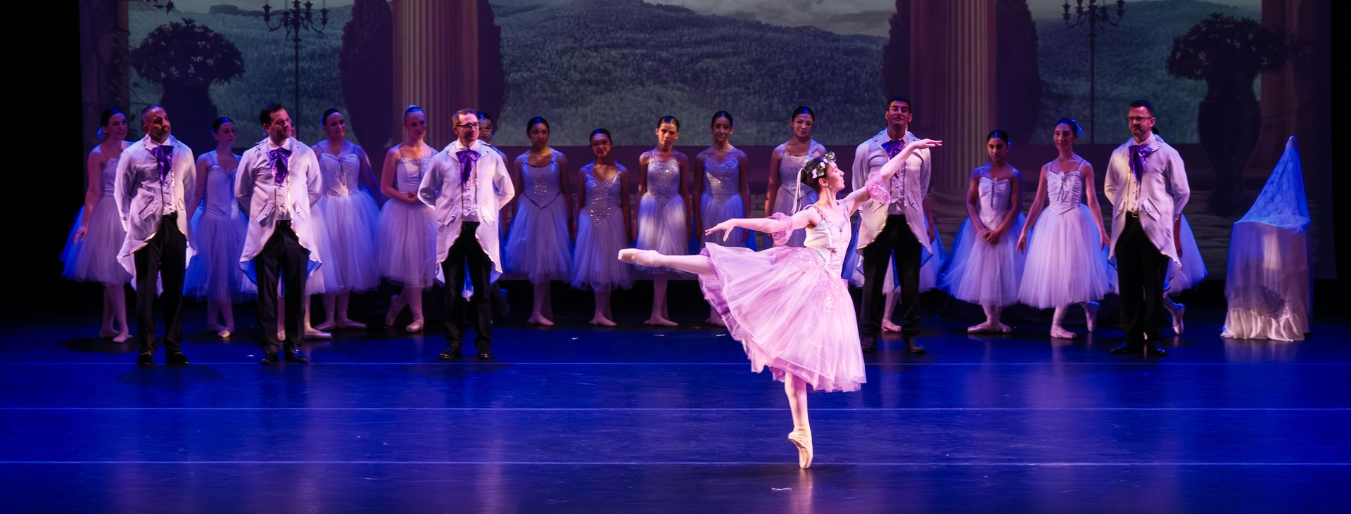 Ballet troupe on stage, with a solo dancer in white posing center under blue-purple lighting.