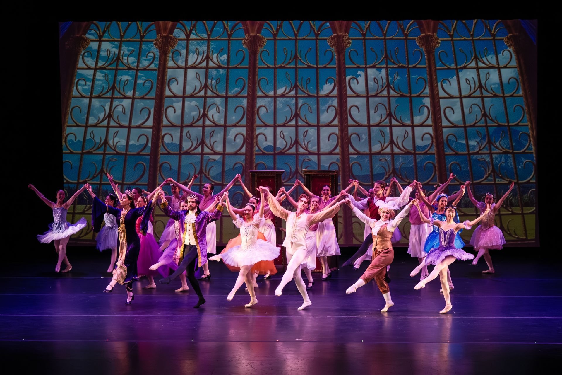 Ballet dancers in colorful costumes perform on a stage before a stained-glass backdrop