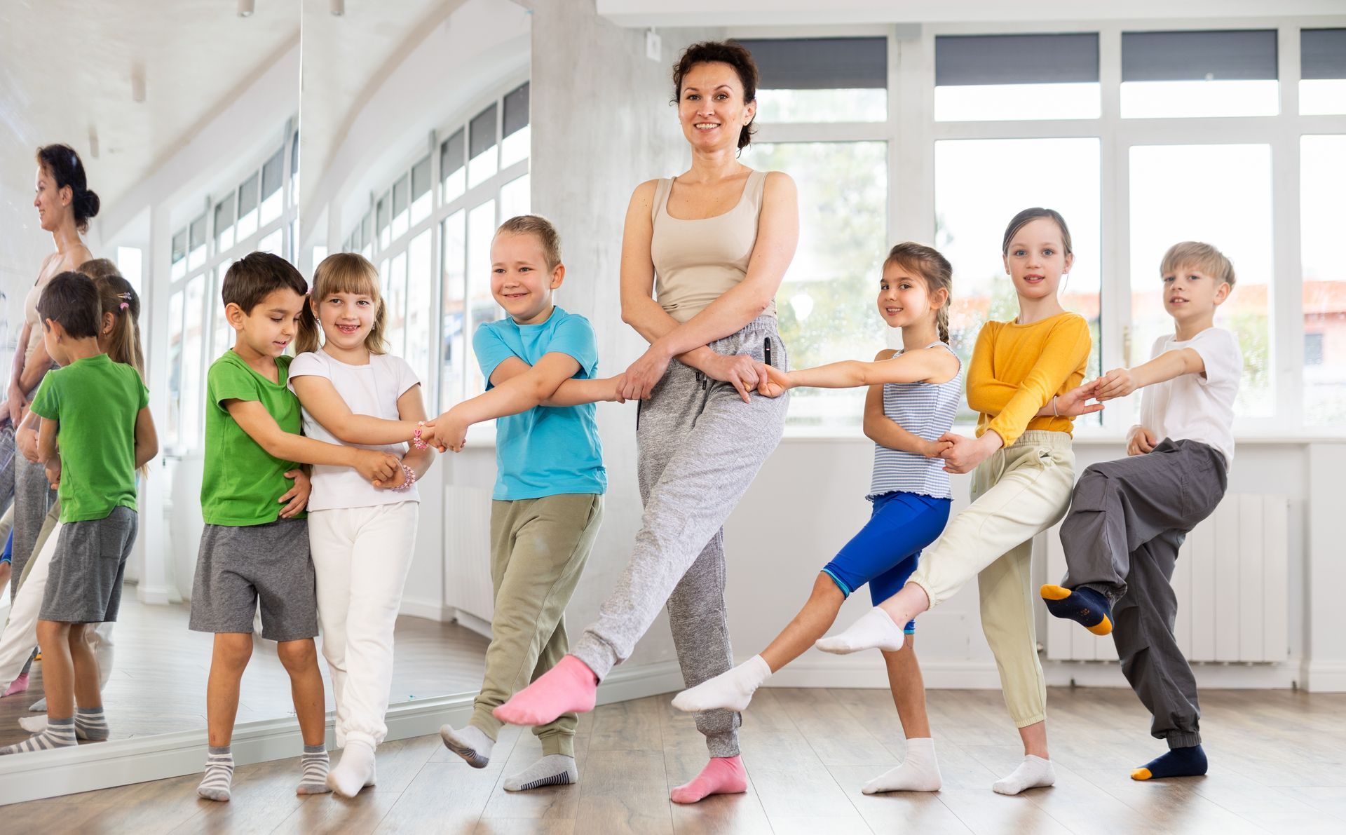 Instructor and children hold hands in a sunlit studio, each extending one leg outward in a stretch.