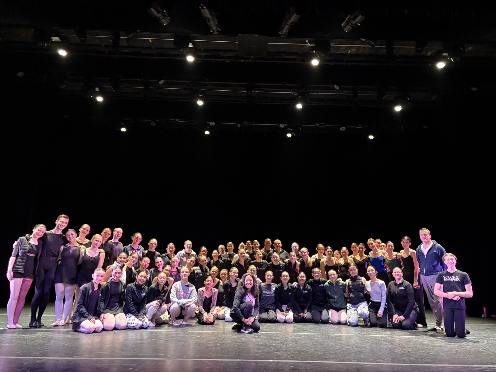 Large group posing on a stage under bright lights, with a black backdrop and many people crouching and standing in rows.