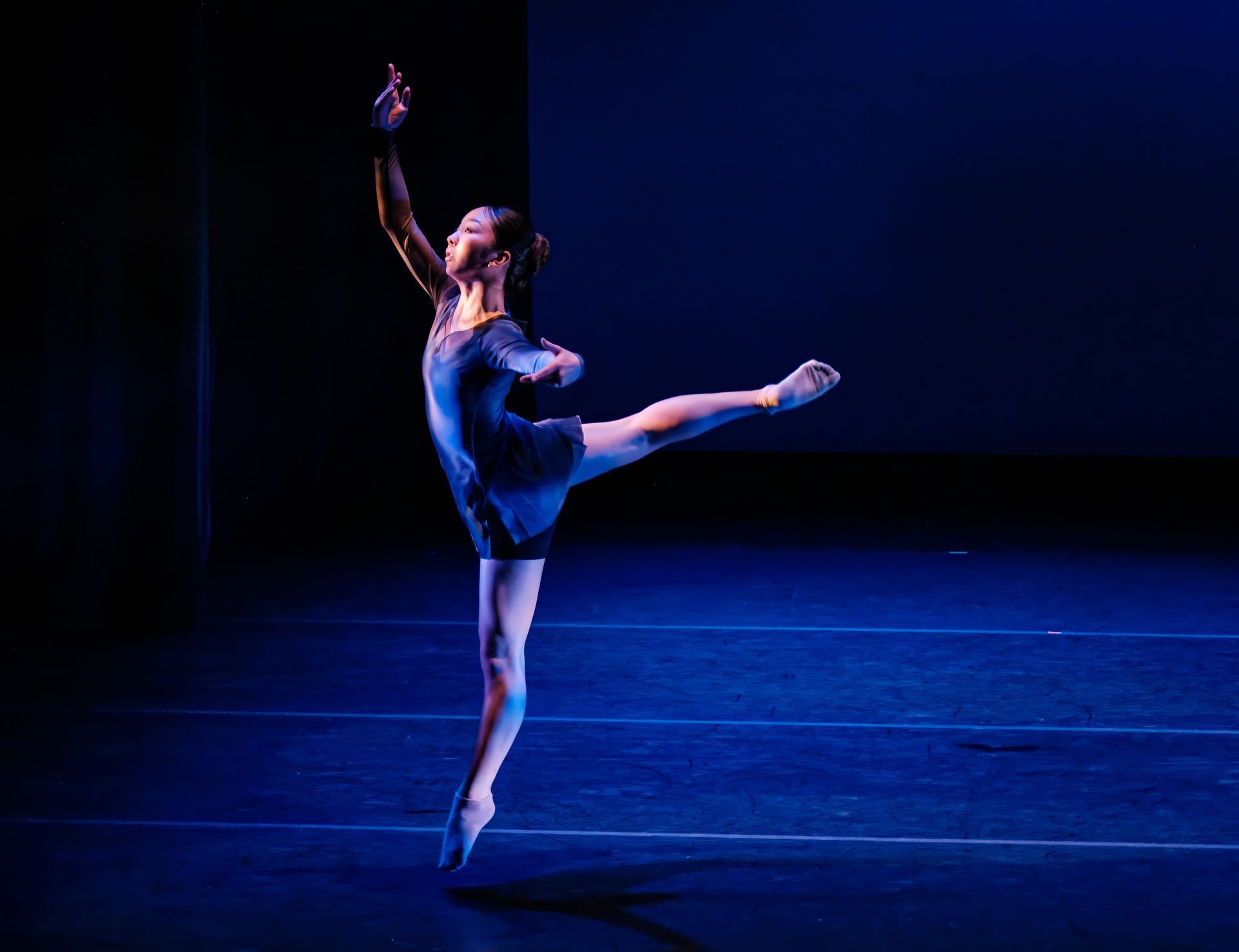 Ballet dancer in blue leotard performing a high leg extension on a dark stage under blue lighting