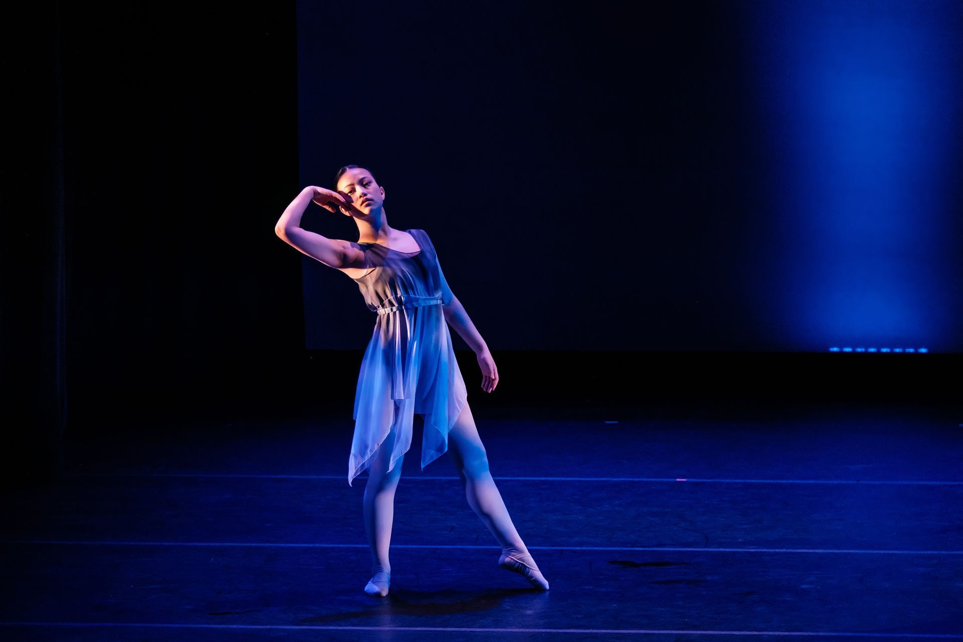 Dancer in a blue dress poses on a dark stage under blue lighting