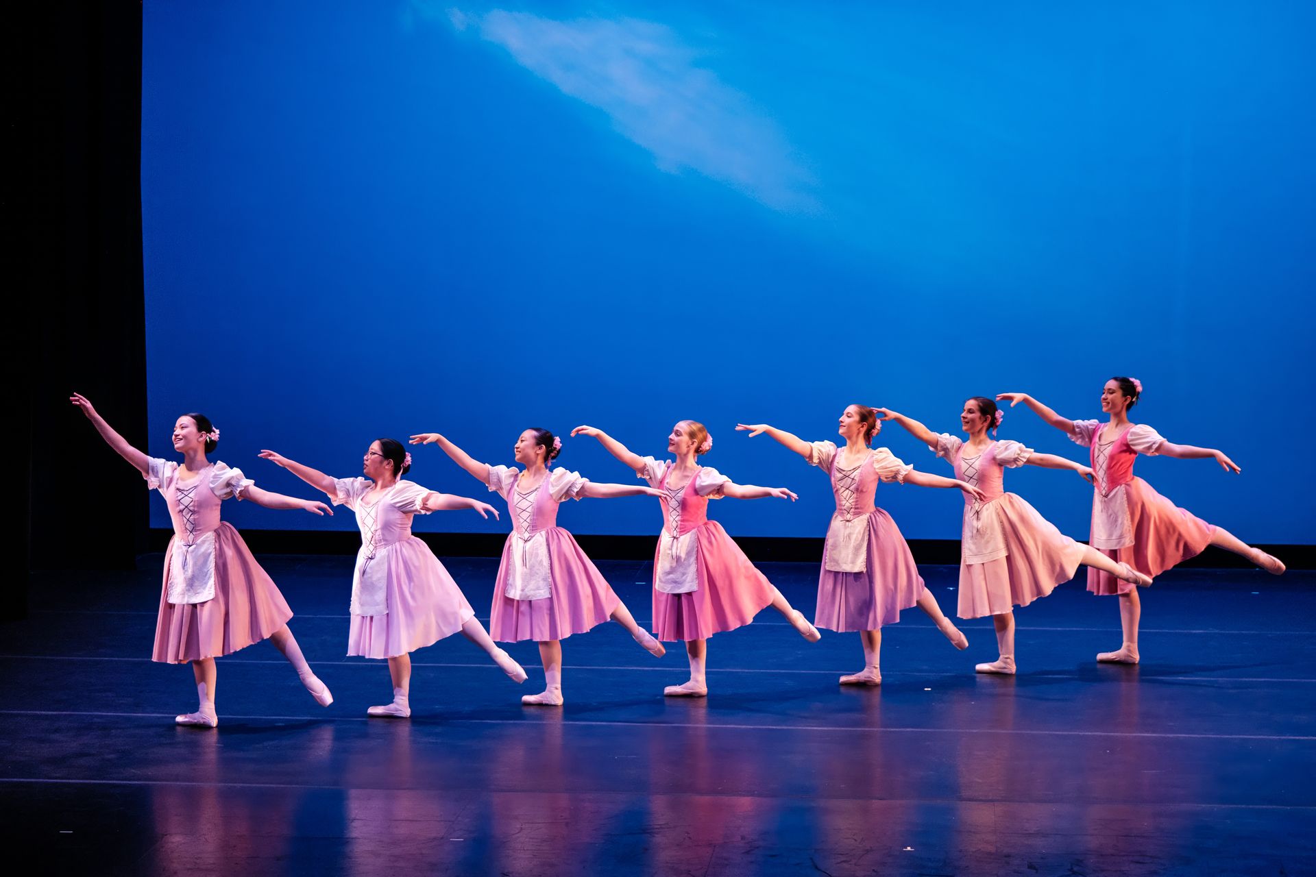 Seven ballet dancers in pink dresses pose in a line on a blue-lit stage