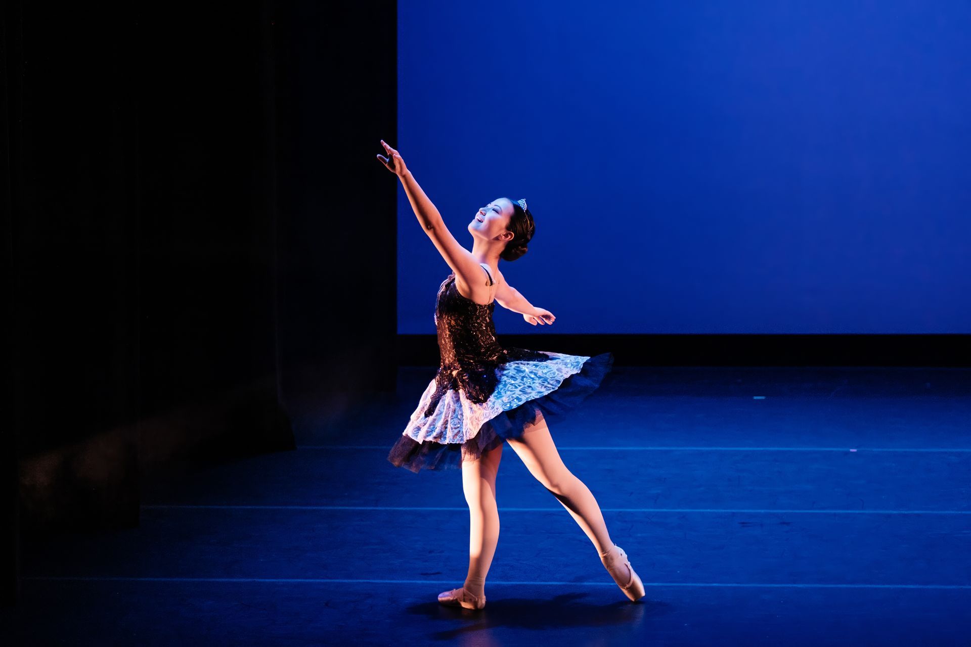 Ballerina in a black-and-white tutu dancing under blue stage lights with one arm raised.