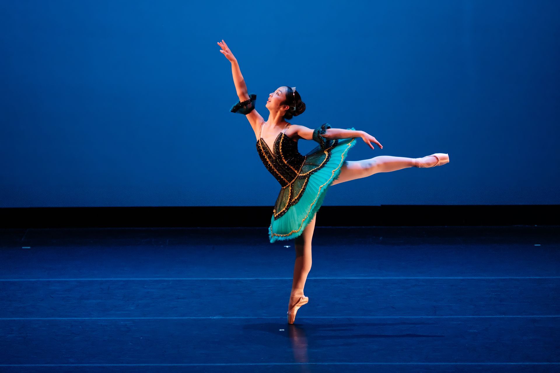 Ballet dancer in turquoise costume balancing on one leg against a blue stage backdrop