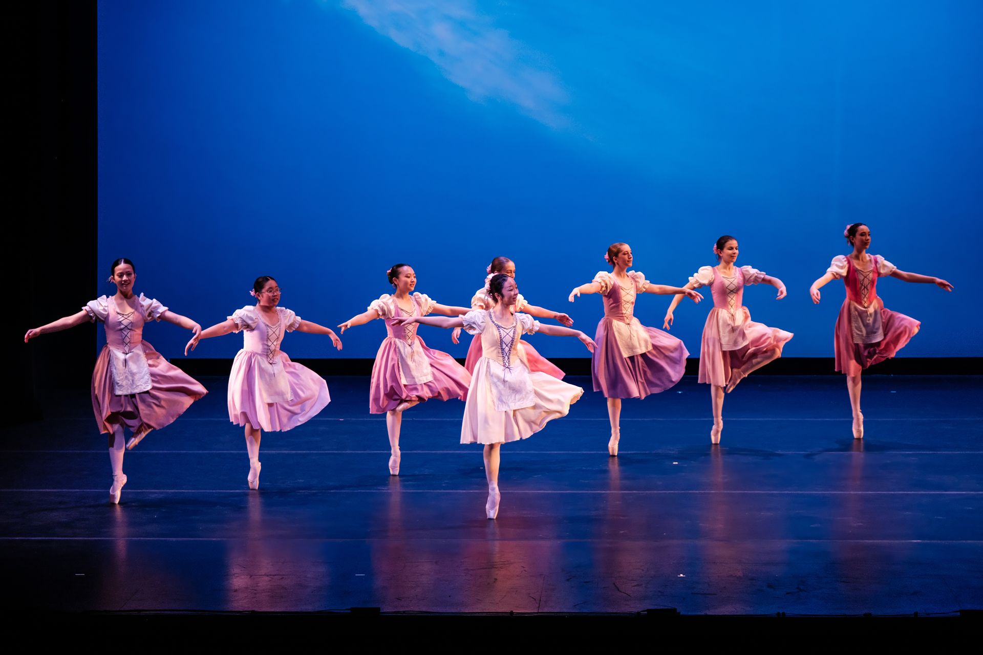 Ballet dancers in pink dresses performing synchronized poses on a blue-lit stage