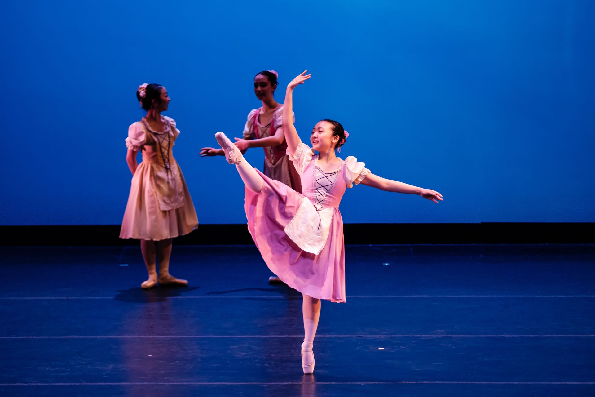 Three ballet dancers in pink costumes pose and dance on a blue-lit stage