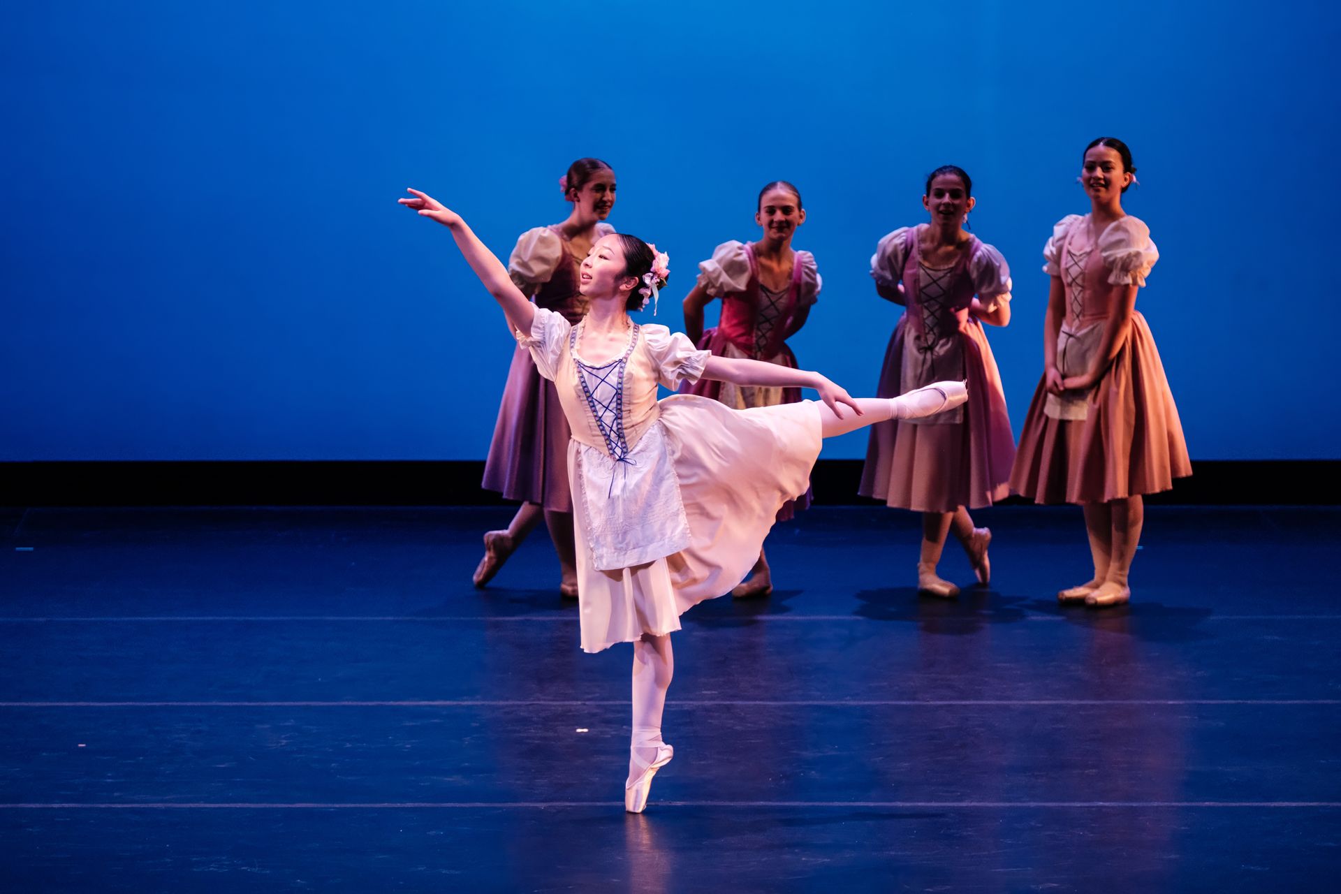 Ballet dancers on a blue-lit stage, with one dancer in a white dress posed center front.
