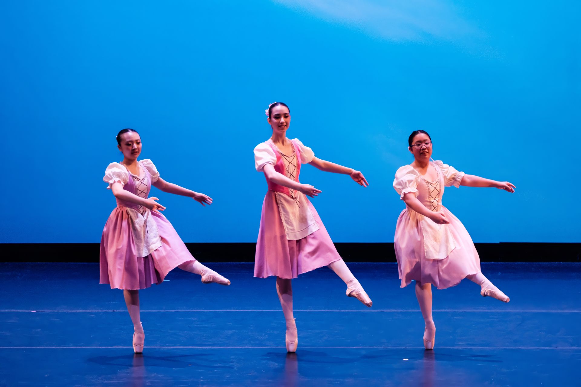Three ballet dancers in pink dresses pose on a blue-lit stage, each balancing with one leg raised.