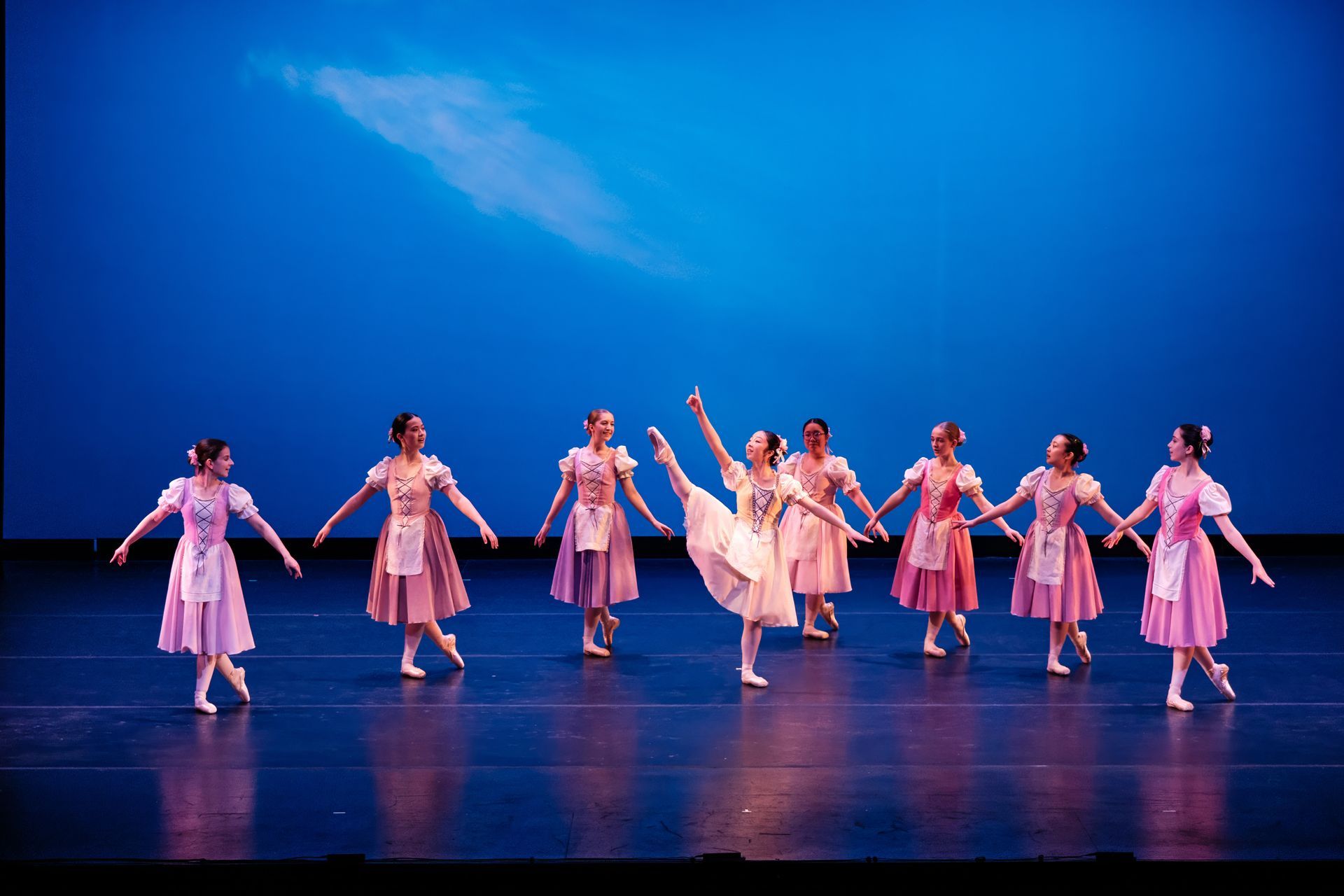 Dancers in pink costumes perform on a blue-lit stage with arms raised